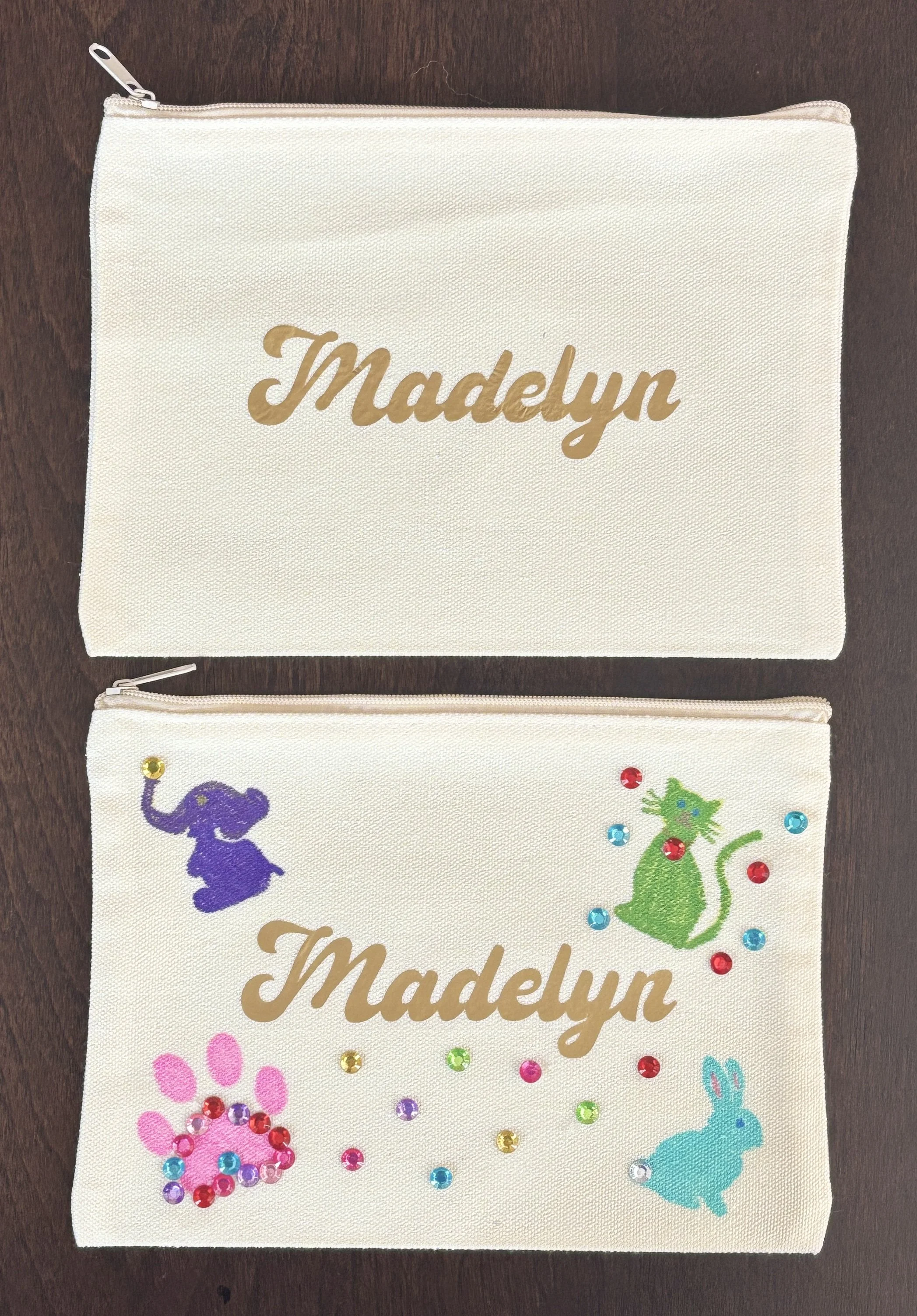 Two cream-colored fabric pouches, each with zippers and decorated with colorful rhinestones and animal silhouettes. The upper pouch has the name "Madelyn" in gold script on it, along with a purple elephant silhouette. The lower pouch features a pink 