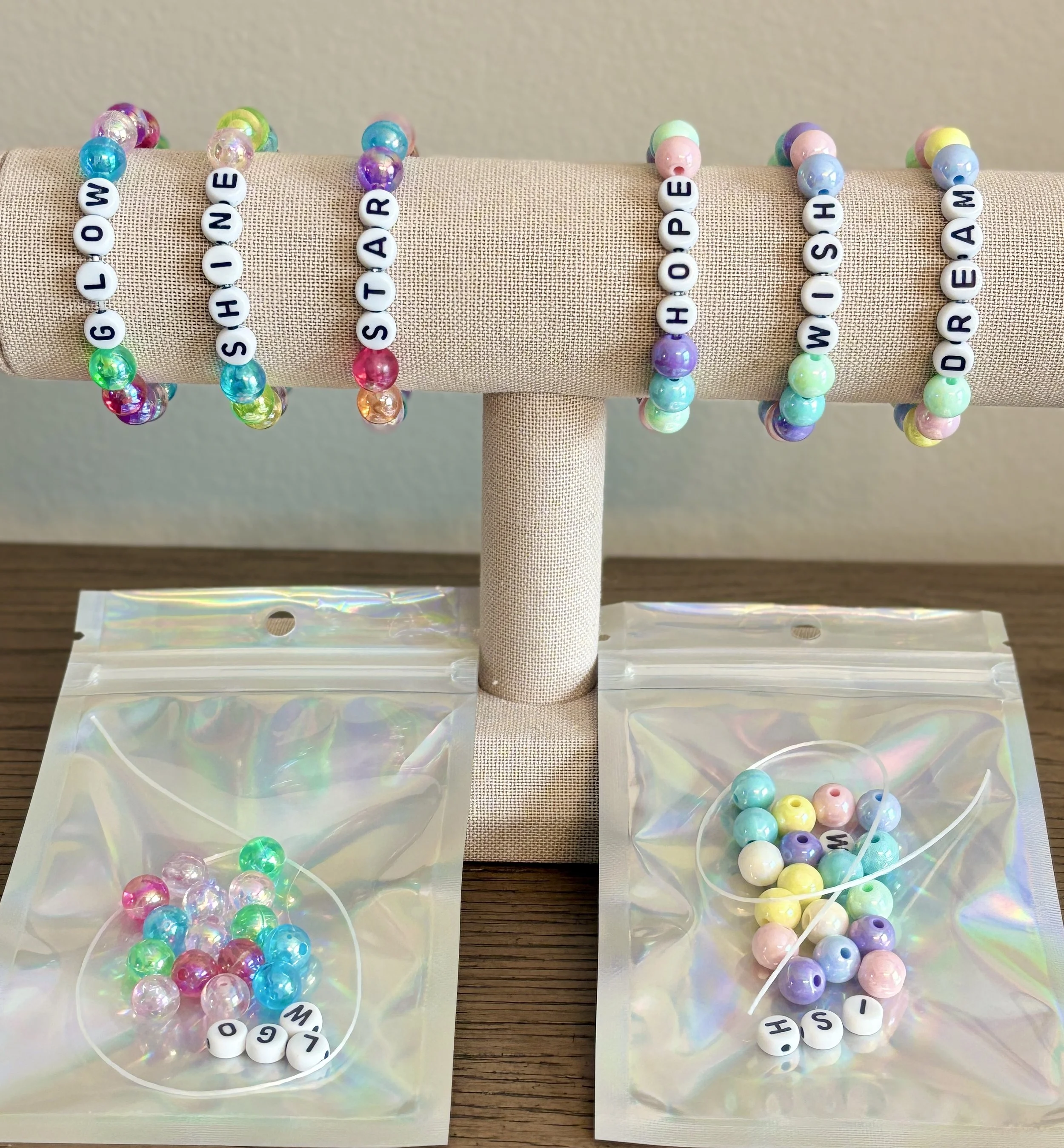 Beaded bracelets spelling out words and names, with colorful beads on a beige fabric display, with some beads free in plastic bags and others with letter beads spelling 'HELLO,' 'SHARE,' 'HOPE,' 'WISH,' and 'DREAM'.
