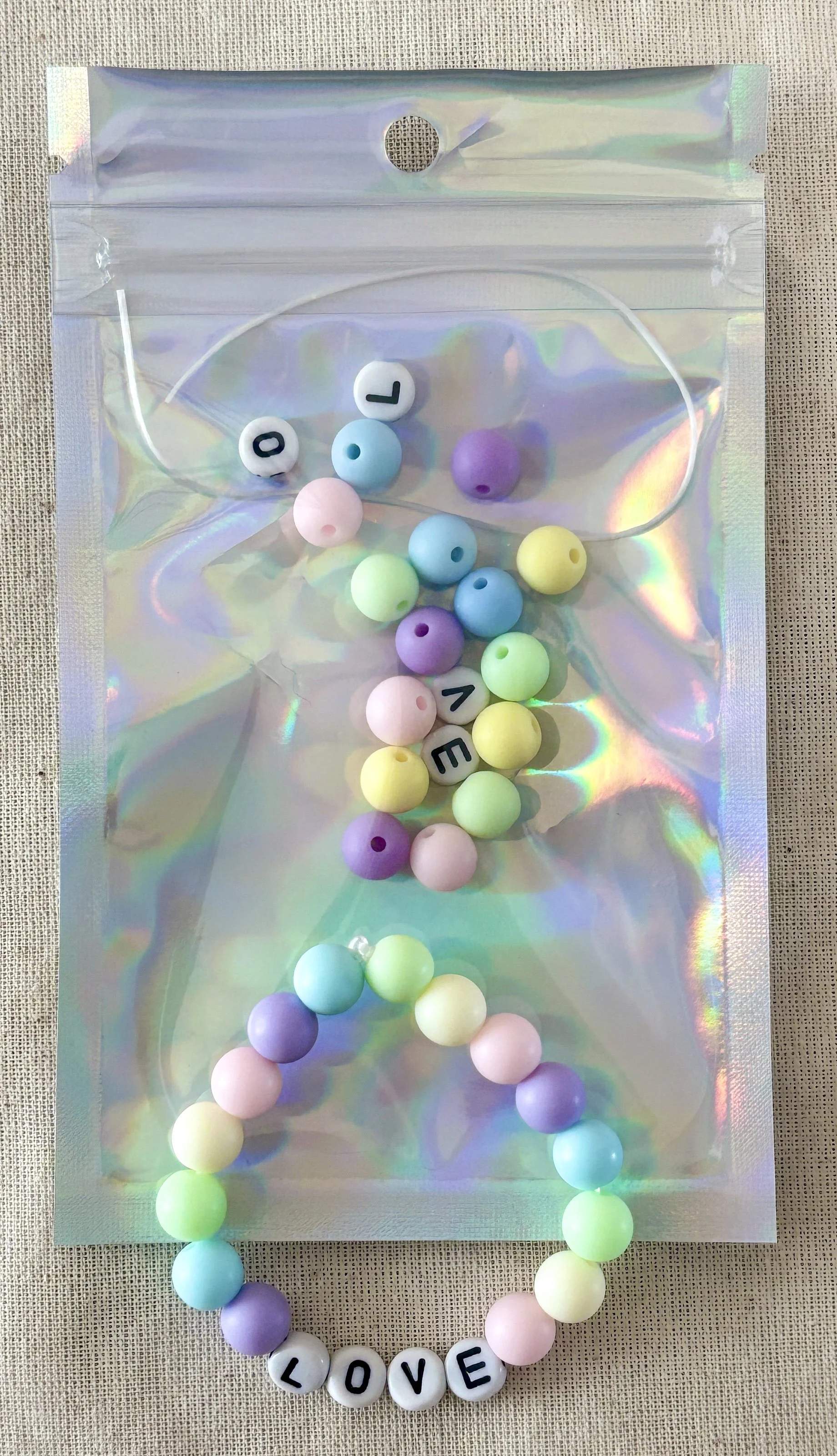 Colorful beaded necklace with the word "LOVE" spelled out with letter beads, placed on holographic packaging on a textured surface.
