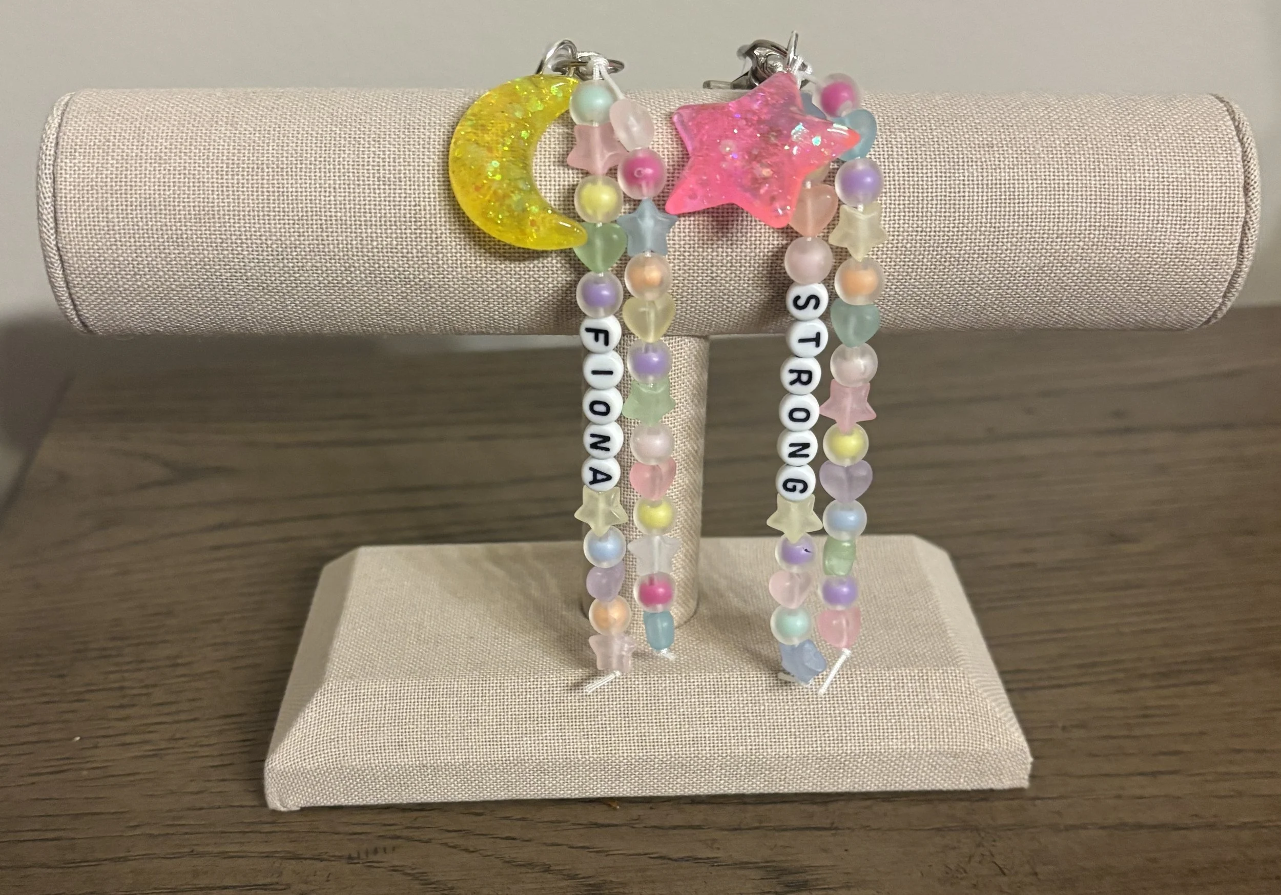 Colorful beaded necklaces and bracelets with star, moon, and letter charms displayed on a beige jewelry stand.