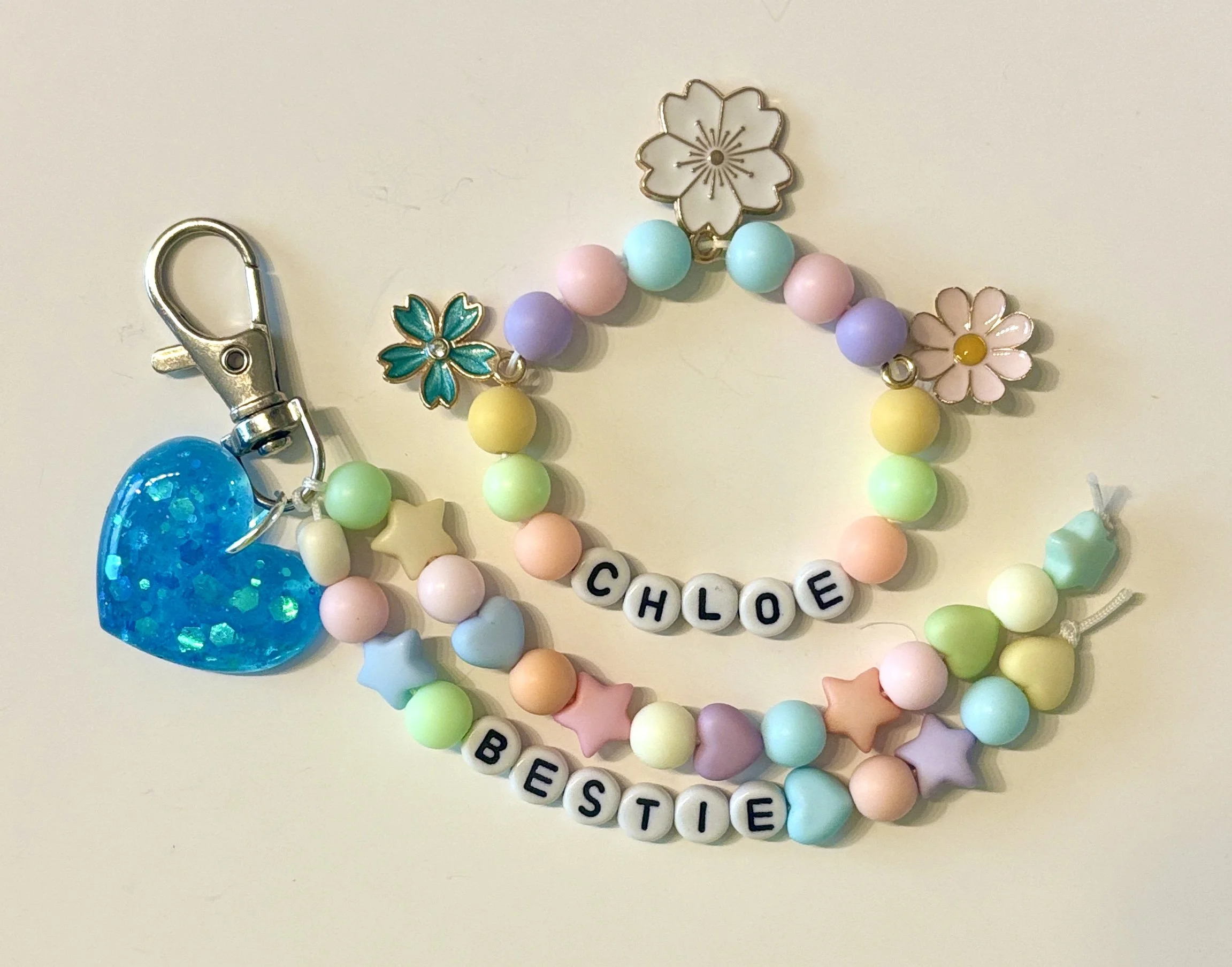 Colorful beaded jewelry with charms, including a blue glittery heart, a gold cherry blossom, a pink flower, and letter beads spelling "BEAUTIFUL".