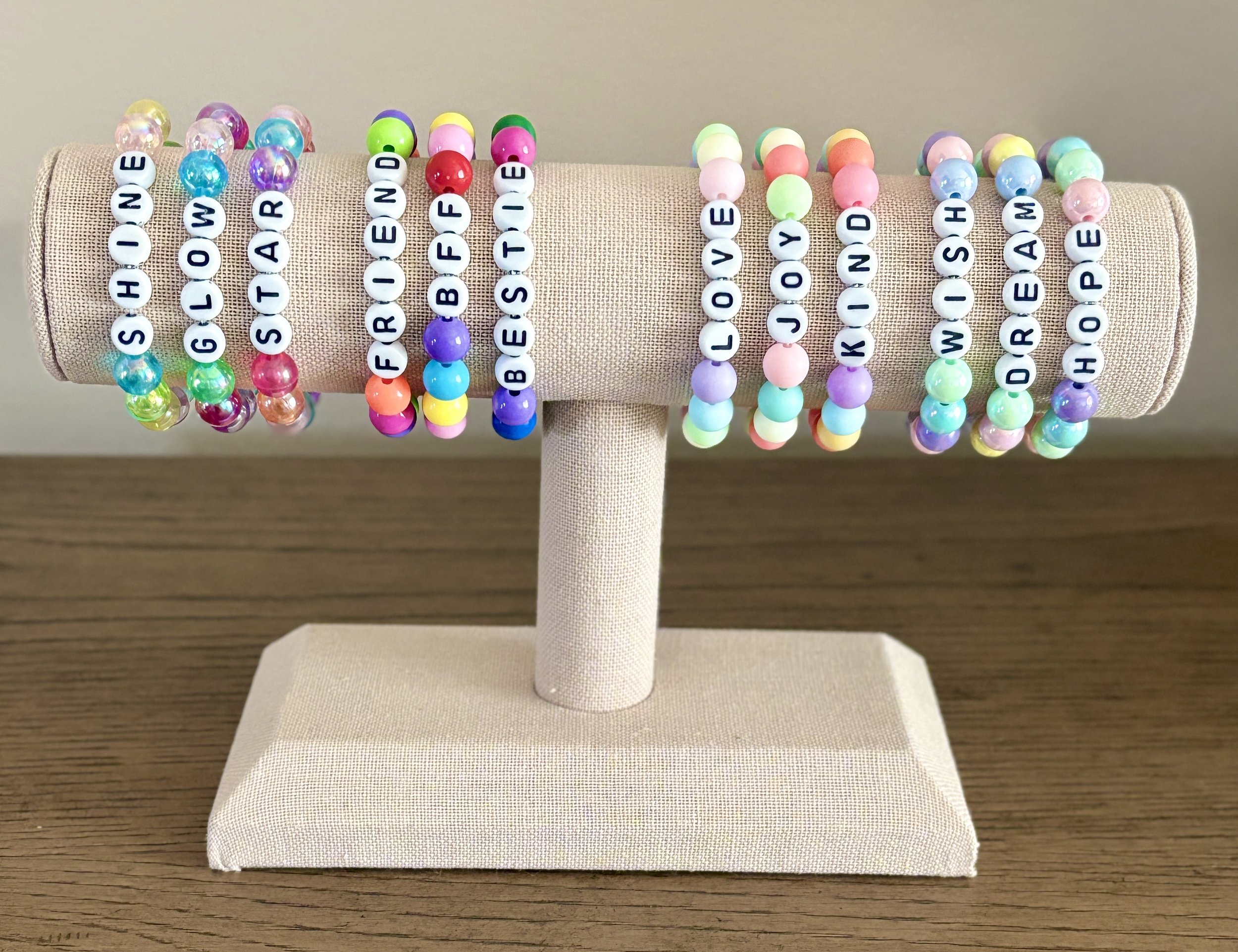 Colorful beaded bracelets displayed on a beige fabric-covered stand with the alphabet beads spelling 'SHOWN', 'GLOAD', 'TAT', 'FEE', 'BIEF', 'SIEBE', 'LOVE', 'YOM', and 'HUSH'.”