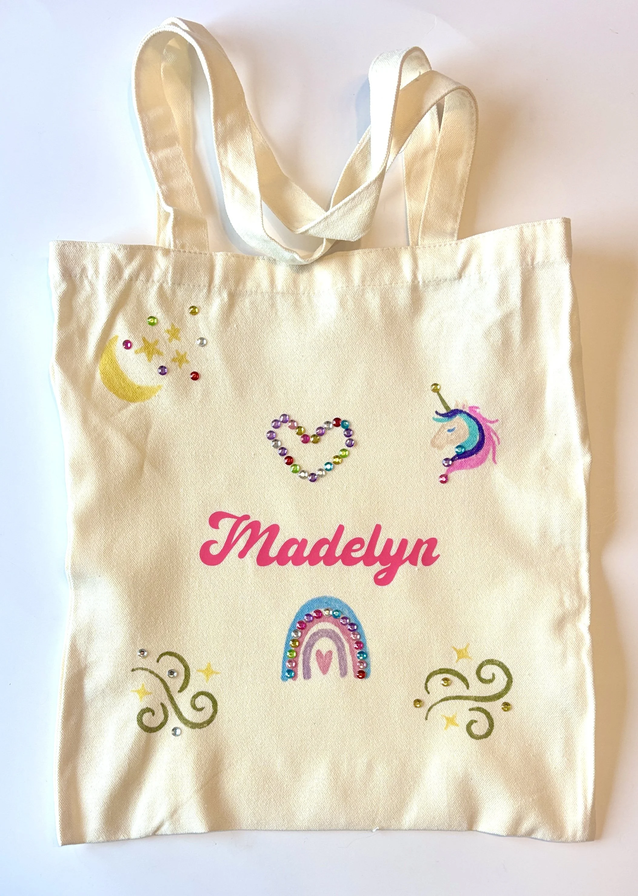 Beige tote bag with colorful unicorn, rainbow, moon, and decorative elements, and the name 'Madelyn' printed in pink.