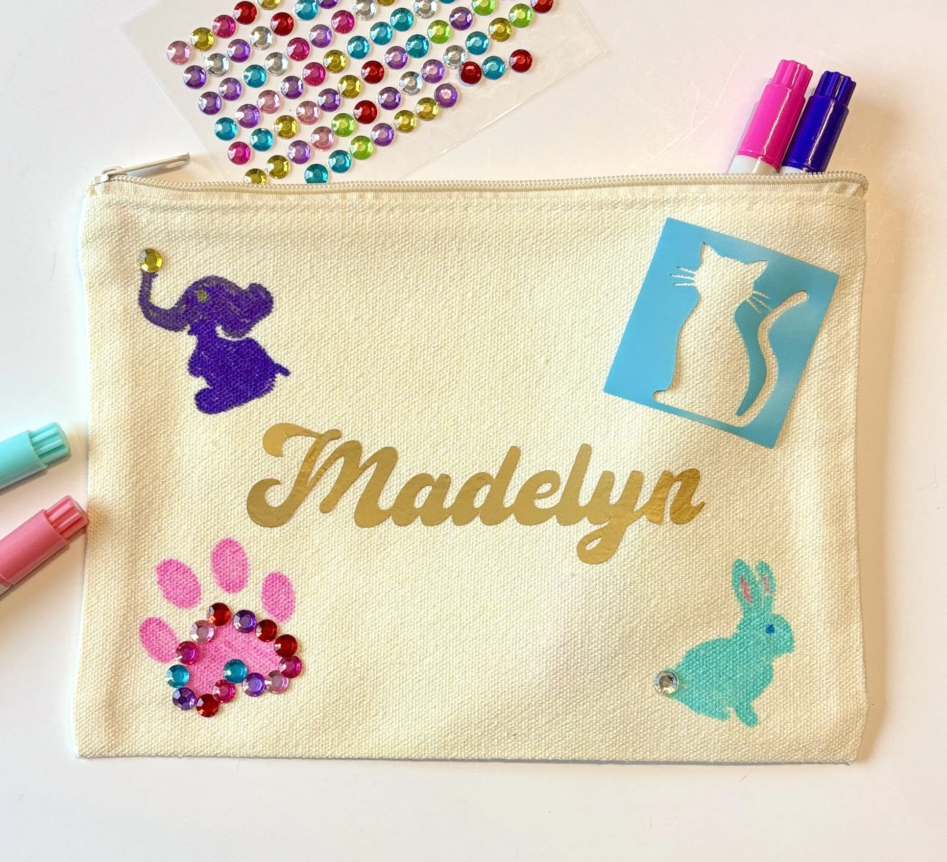 A beige fabric pencil case with a purple embroidered elephant, gold cursive text, and various colorful stickers and embellishments, including rhinestones, pom-pom designs, and markers, on a white background.