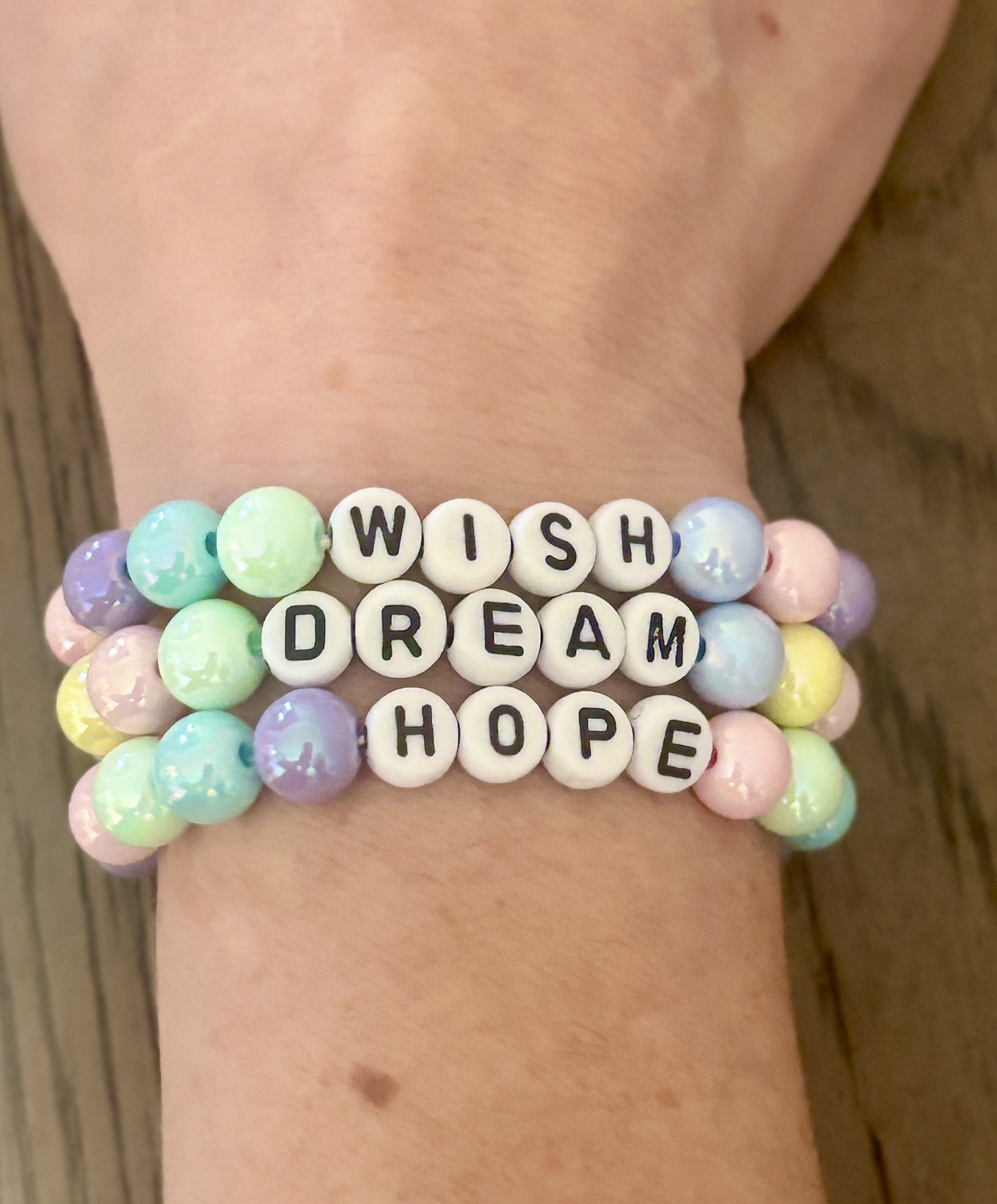 A bracelet of pastel-colored beads with white letter beads spelling out inspirational words: 'WISH,' 'DREAM,' and 'HOPE', worn on a person's wrist.