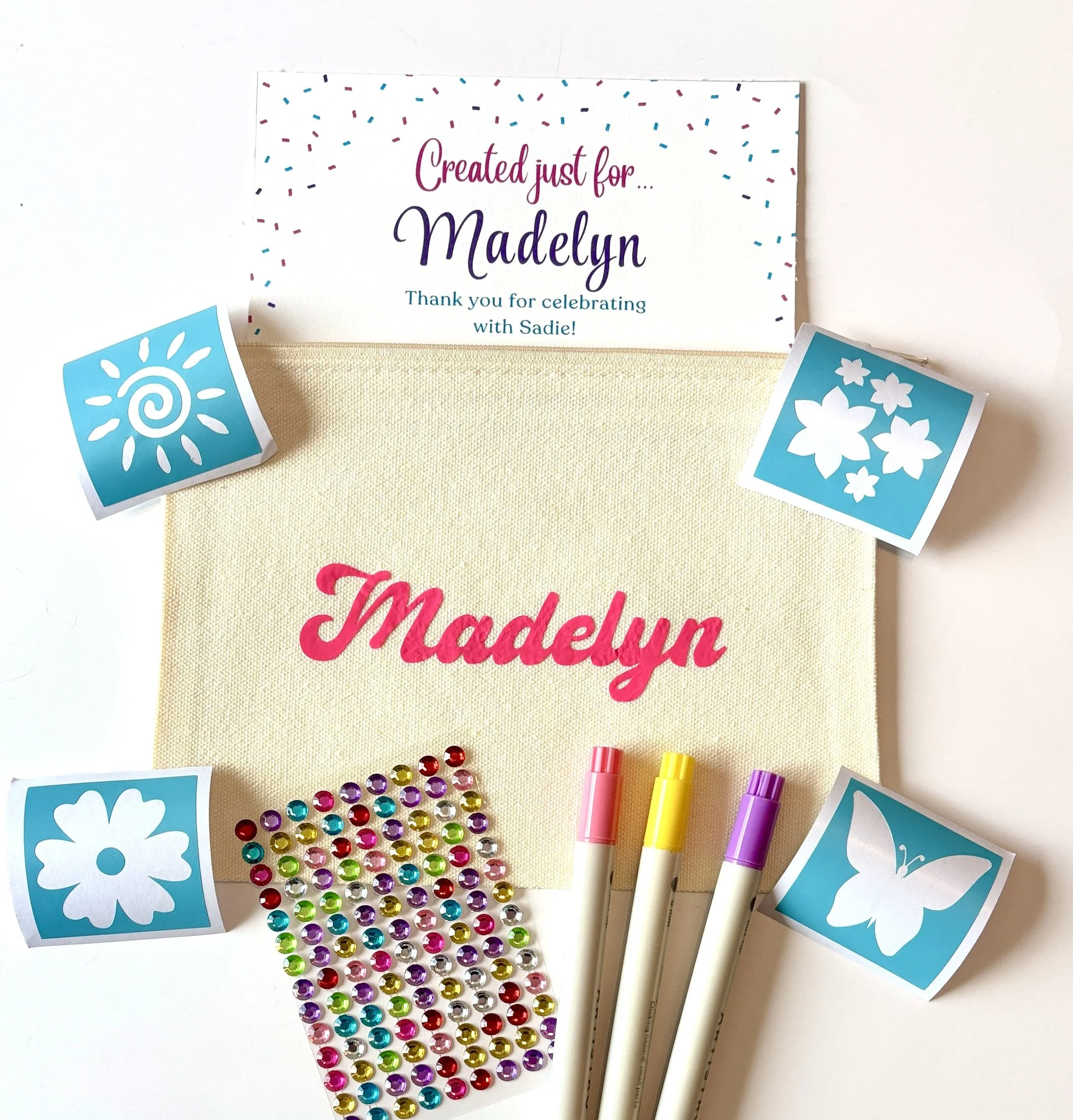 Craft supplies including colorful rhinestones, markers, decorative paper with floral and sun patterns, and a card with the text 'Created just for Madelyn. Thank you for celebrating with Sadie!'