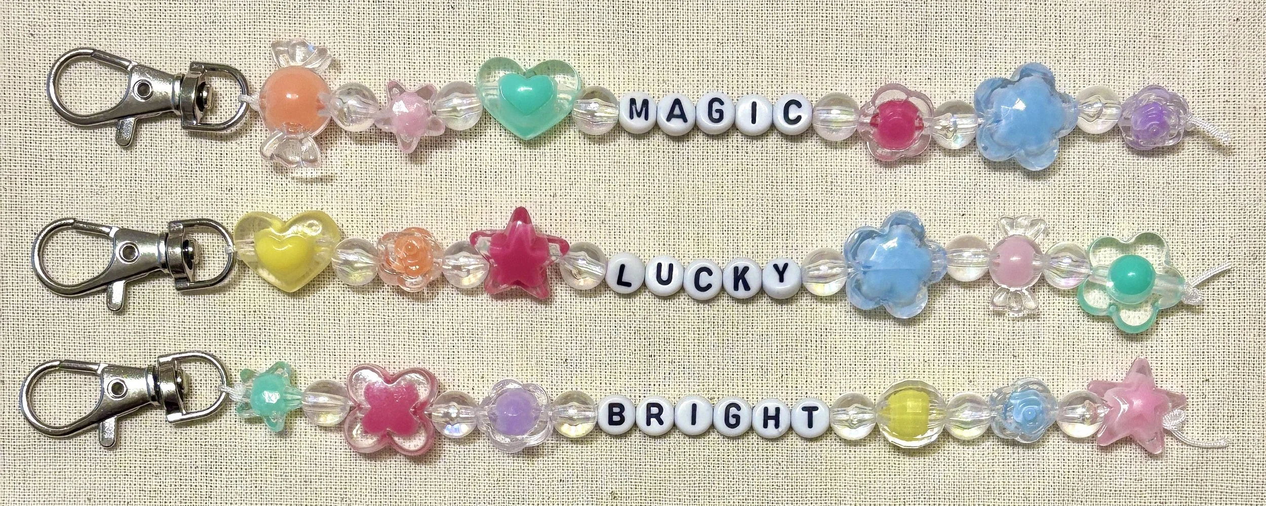 Three beaded keychains featuring colorful flowers, hearts, stars, and beads. Each keychain spells out words with letter beads: 'MAGIC,' 'LUCKY,' and 'BRIGHT.'