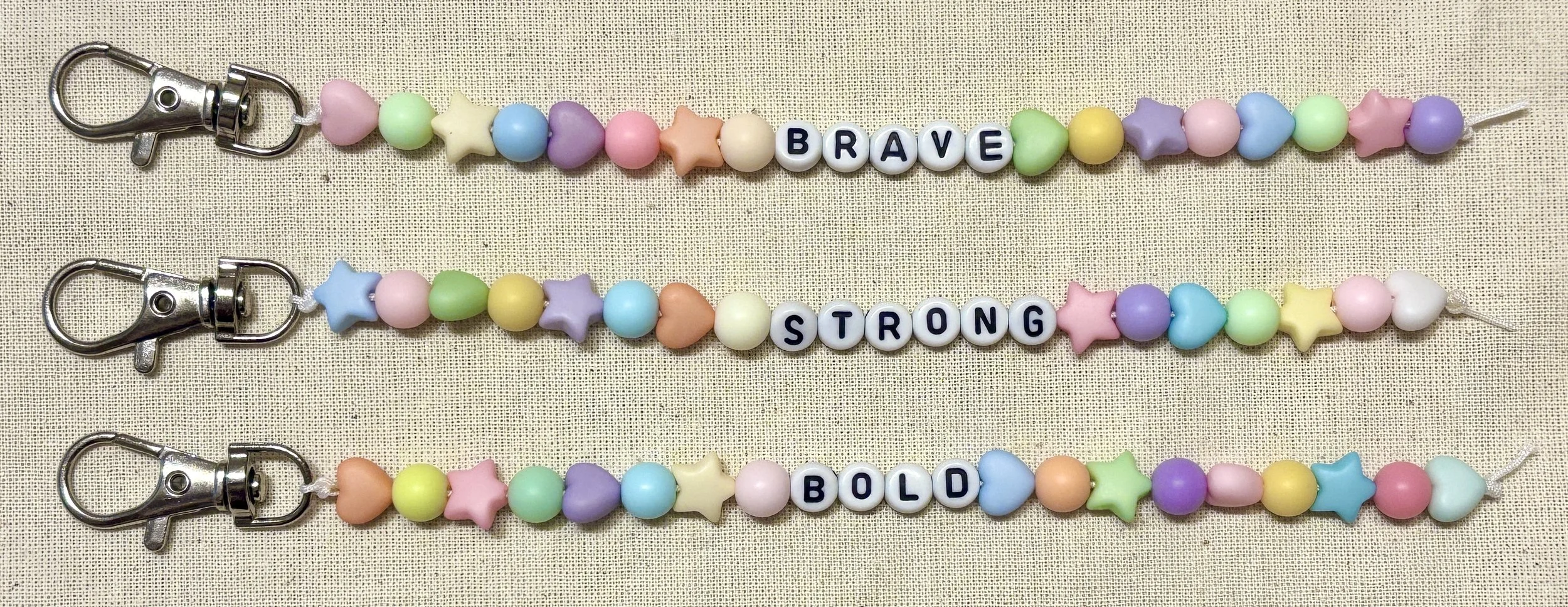 Colorful beaded keychains with star and heart shapes, each spelling out words: 'BRAVE', 'STRONG', and 'BOLD'.