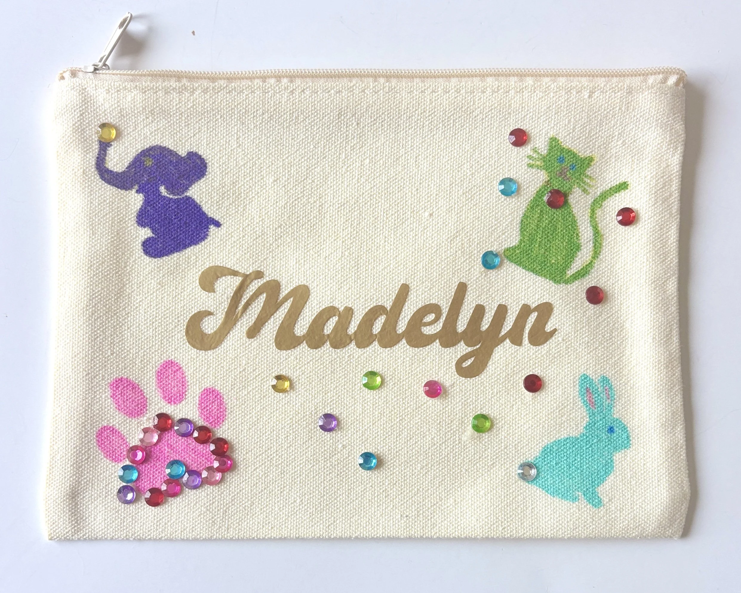 A beige fabric pouch decorated with colorful rhinestones, featuring embroidered purple and green animals, along with pink and blue patterns.