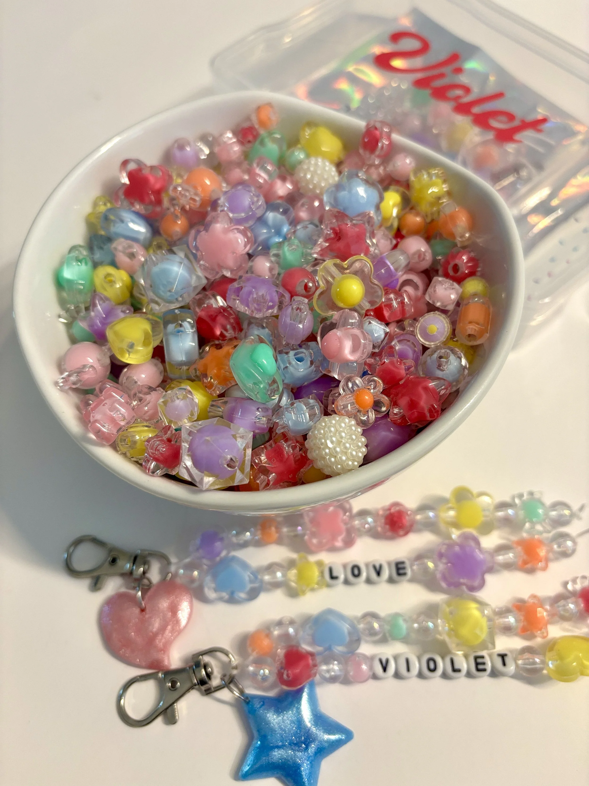 A white heart-shaped bowl filled with multicolored beads and decorative charms, including a pink heart, star-shaped blue bead, and pearl-covered white sphere. There are also letter beads spelling out 'LOVE' and 'VIOLET,' along with a blue star-shaped