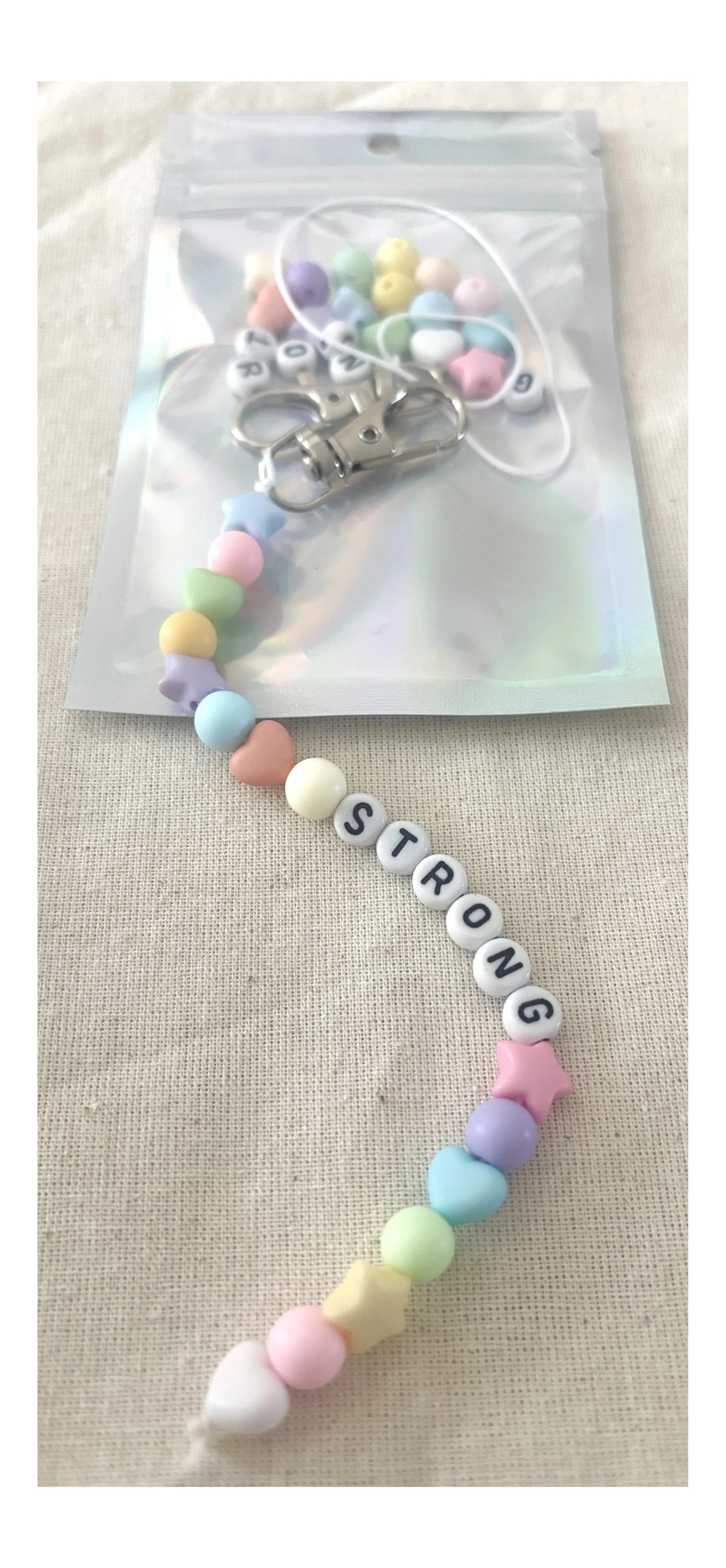 A pastel-colored beaded necklace spelling 'STRONG' with star and heart beads, placed on a beige fabric surface, with a plastic bag containing additional beads and a metal clasp.