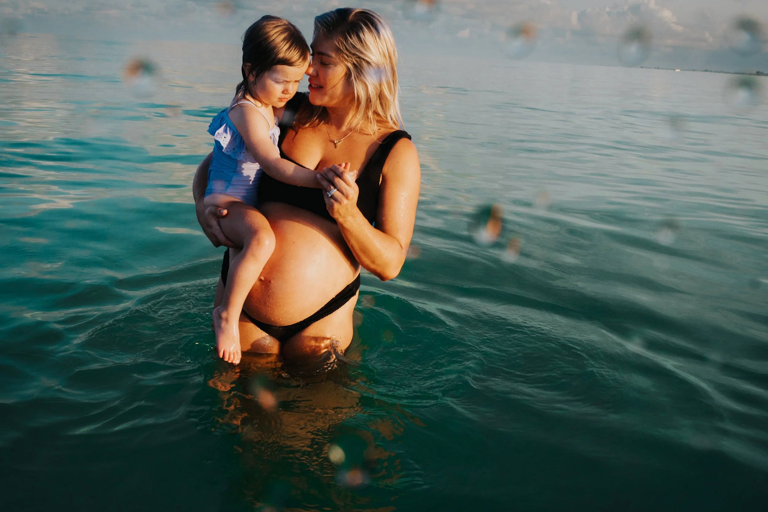 A woman with blonde hair holding a young girl in a blue swimsuit in a body of water, with a calm and scenic backdrop of water and distant land.