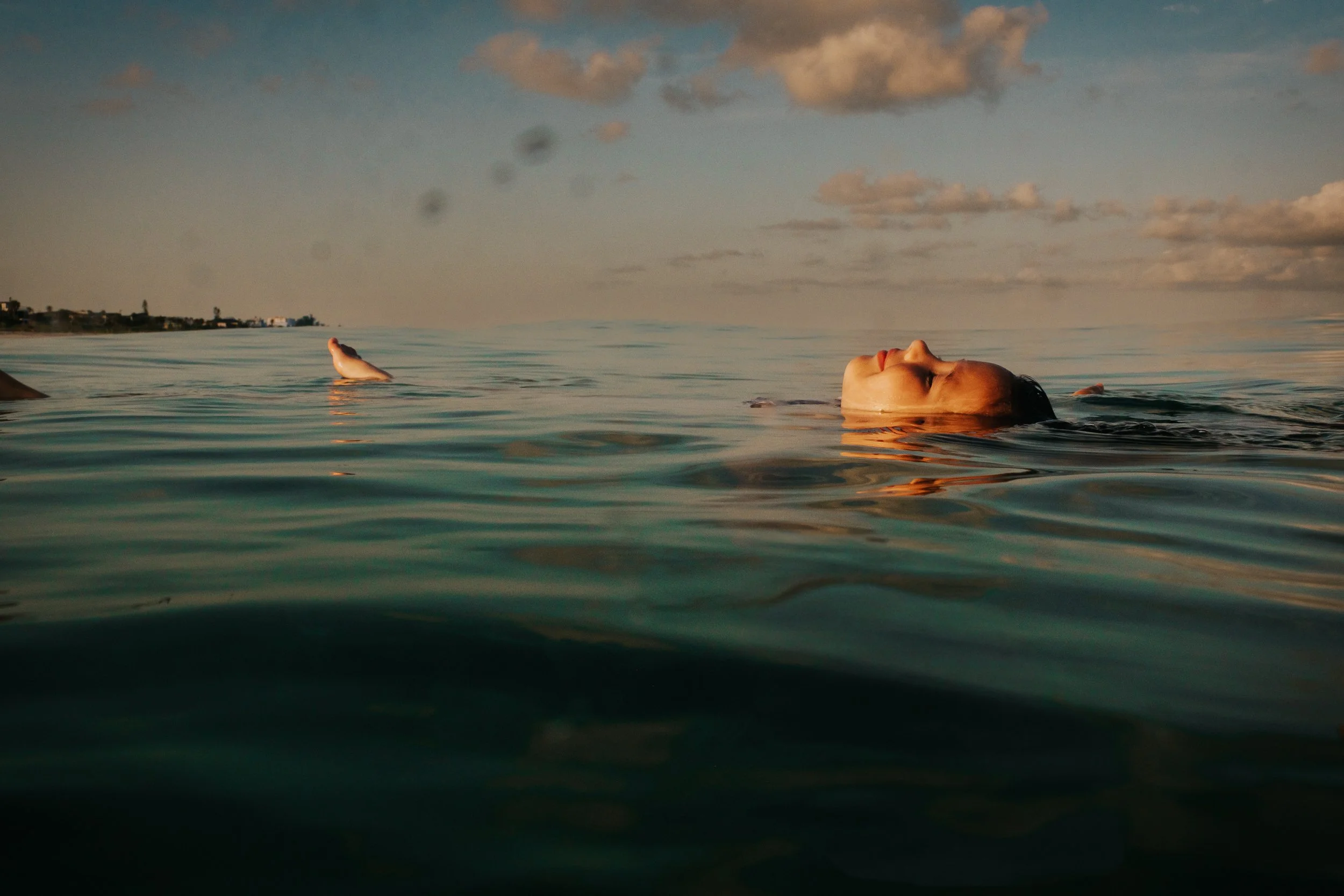 Person floating on back in calm ocean water at sunset with clouds in the sky.