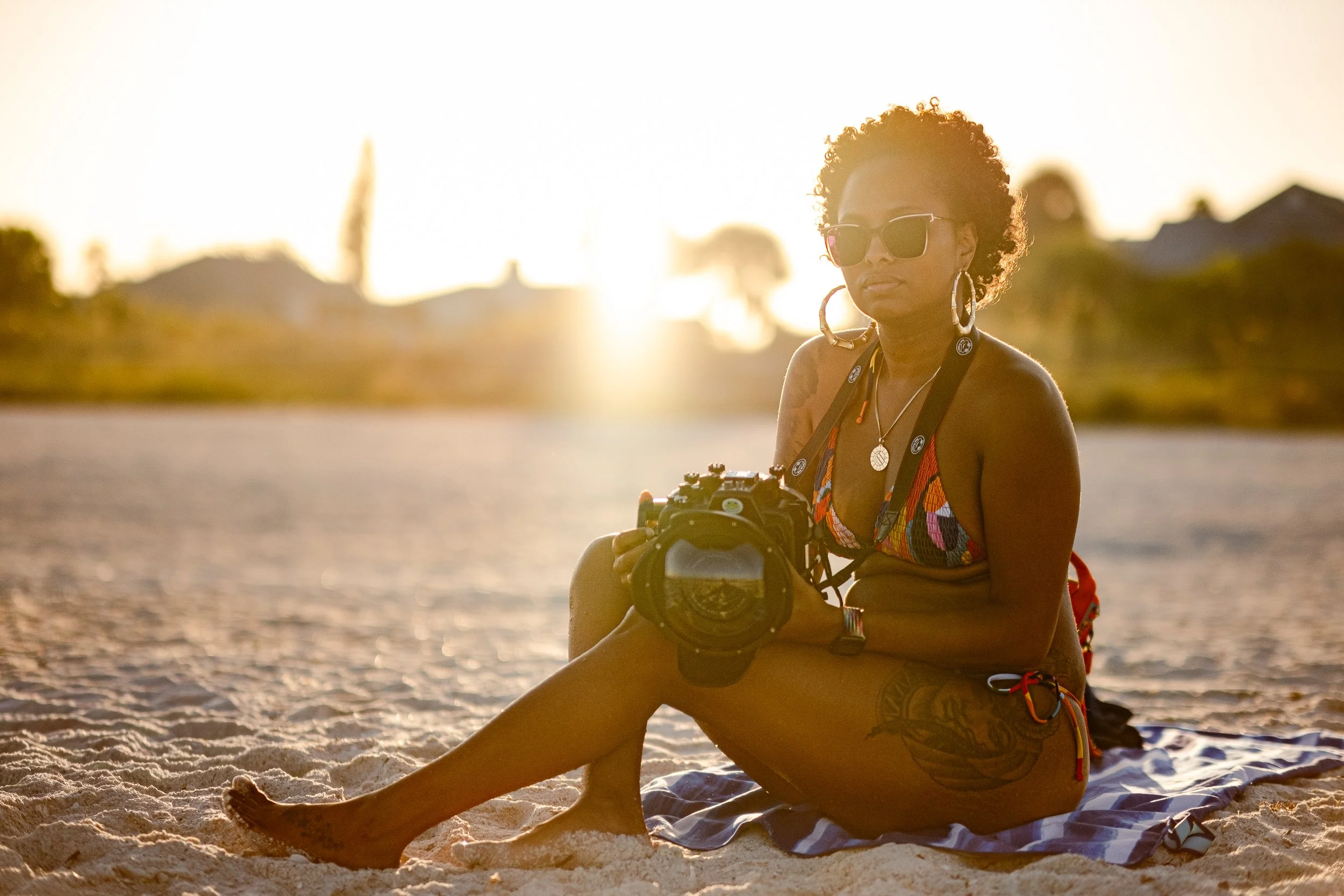 A woman sitting on a sandy beach during sunset, holding a camera in her hands, wearing sunglasses, a colorful patterned swimsuit top, and large hoop earrings.