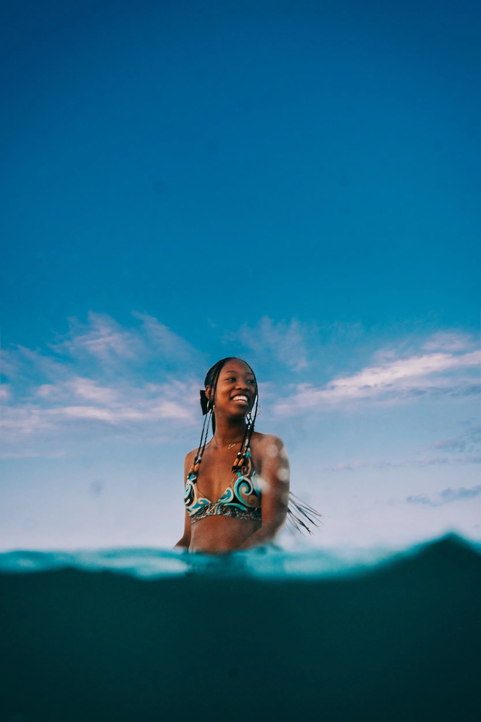 A woman with braided hair wearing a colorful bikini top stands in the water with the ocean and blue sky in the background.
