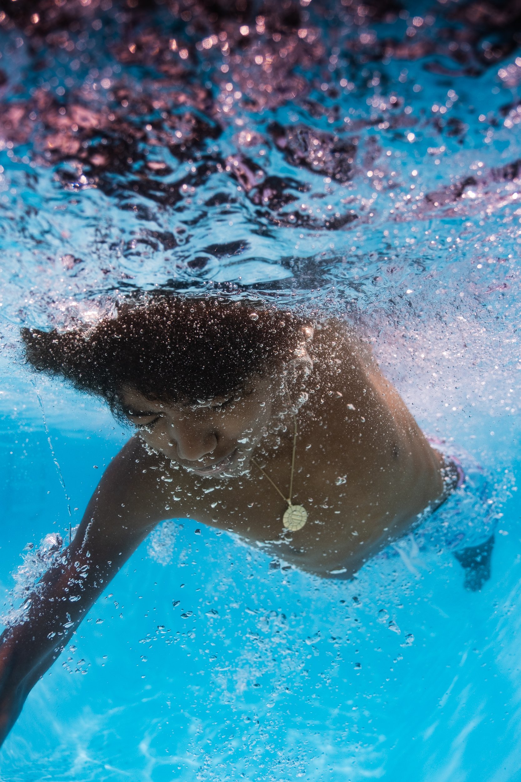A person swimming underwater in a pool, wearing a gold necklace with a round pendant.