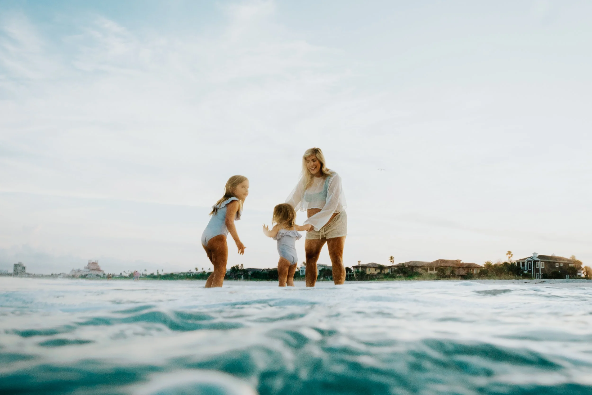 A woman and three young girls playing or wading in the ocean at sunset or late afternoon, with houses and palm trees in the background.