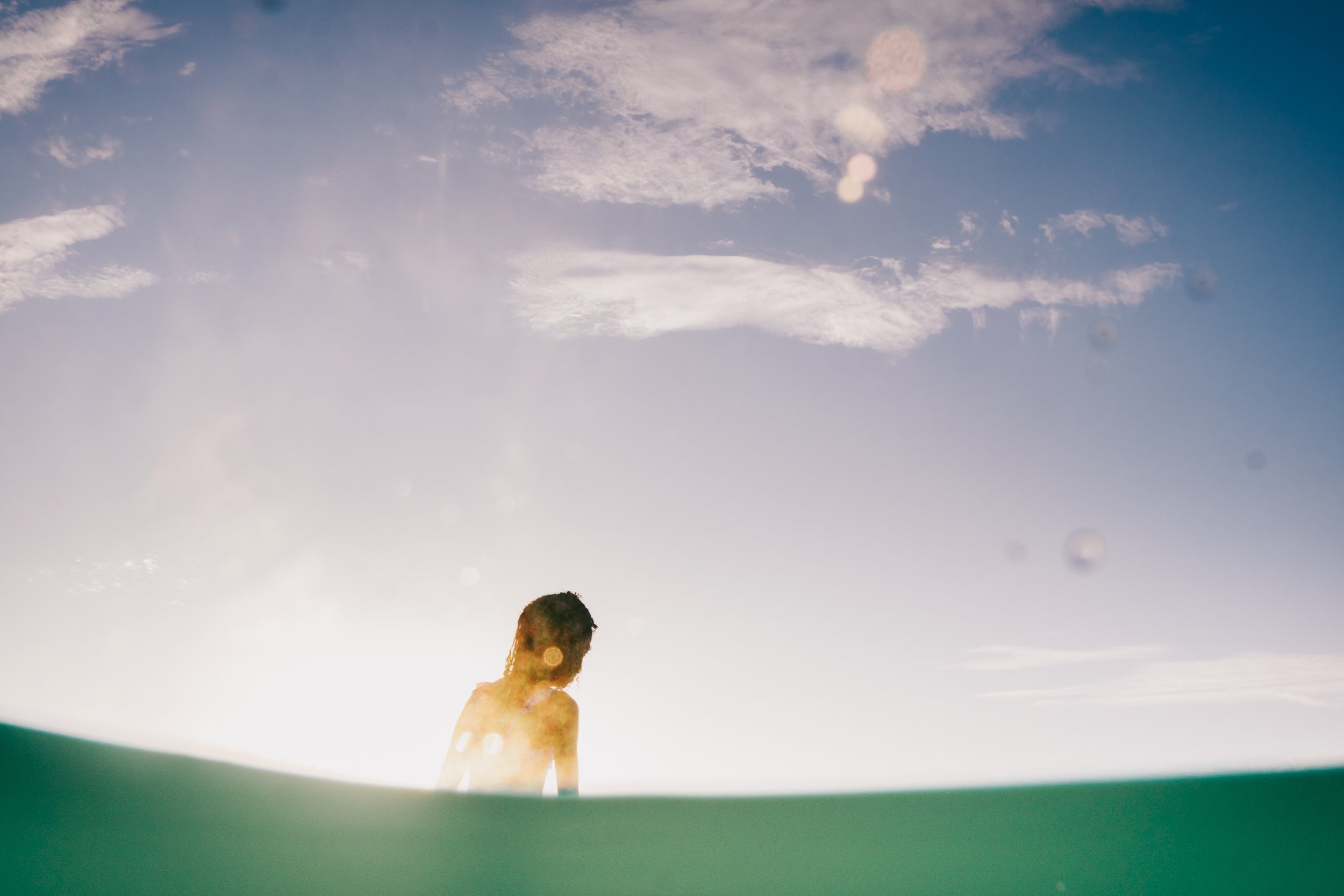 A person with long hair standing outdoors near the water during sunset or sunrise, with the sky partly cloudy and the moon visible.