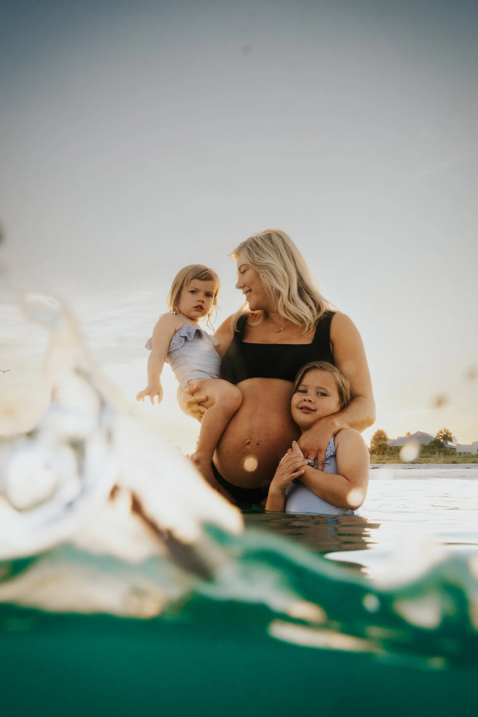 A pregnant woman and two young girls in swimsuits enjoying time by the water, with a boat in the background at sunset.