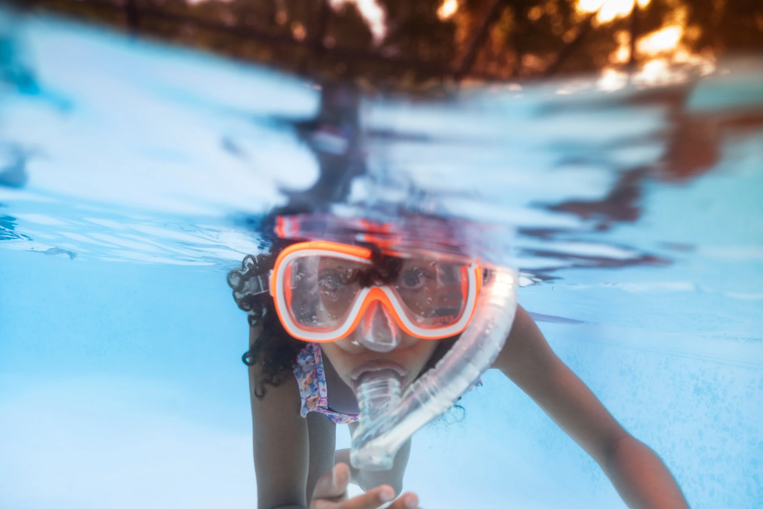 A child swimming underwater in a blue pool, wearing a snorkeling mask and snorkel, with curly hair and a colorful swimsuit.