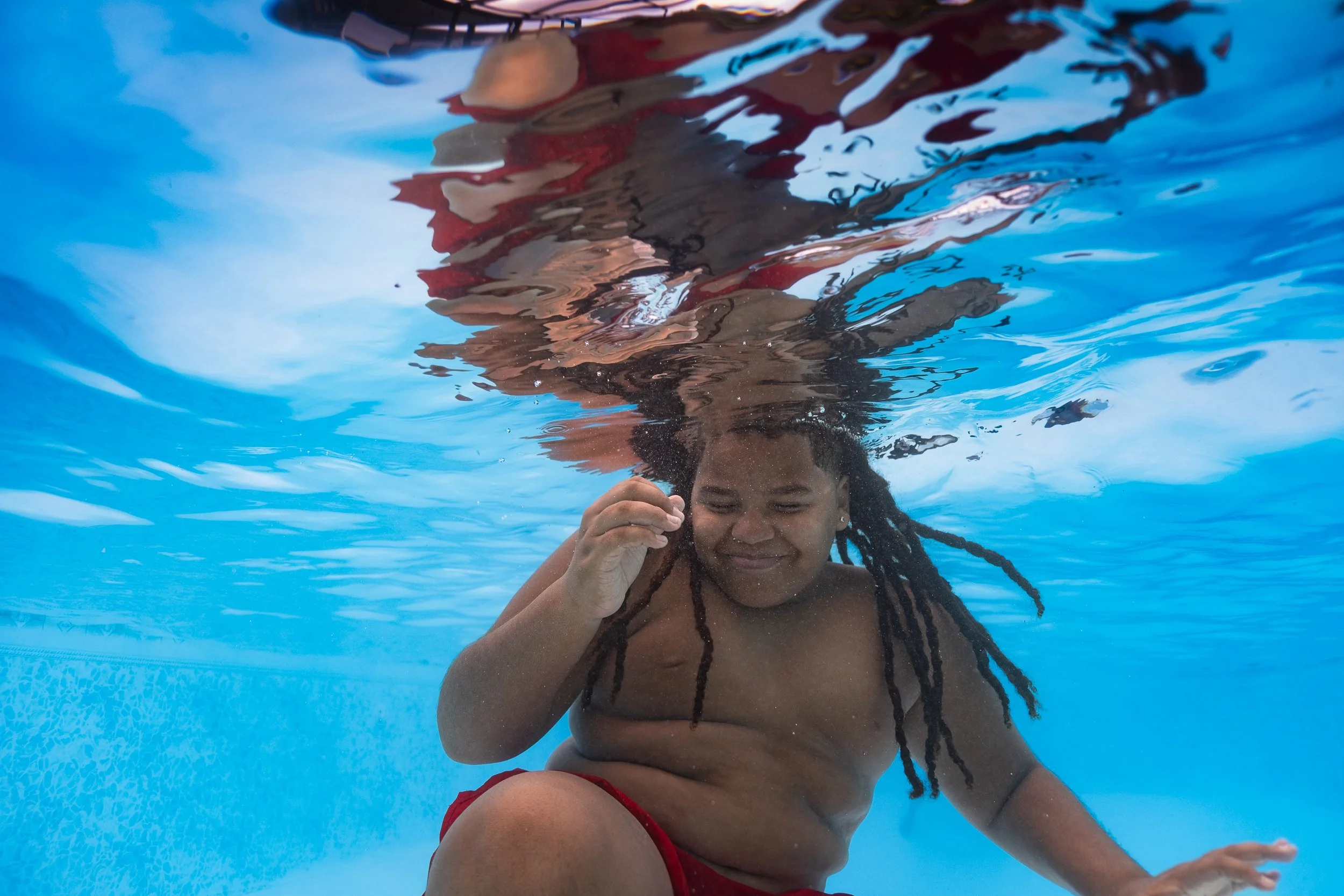 A woman with dreadlocks swimming underwater in a pool, wearing a red swimsuit and smiling.