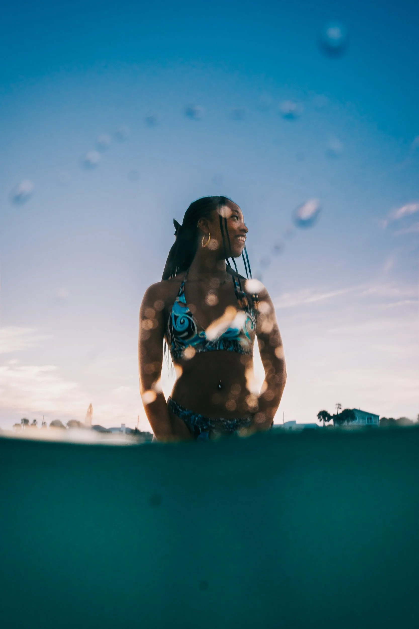 A woman smiling and looking to the right, wearing a blue bikini with black patterns, standing outdoors during sunset with houses and palm trees in the background, and raindrops on a glass or window in the foreground.