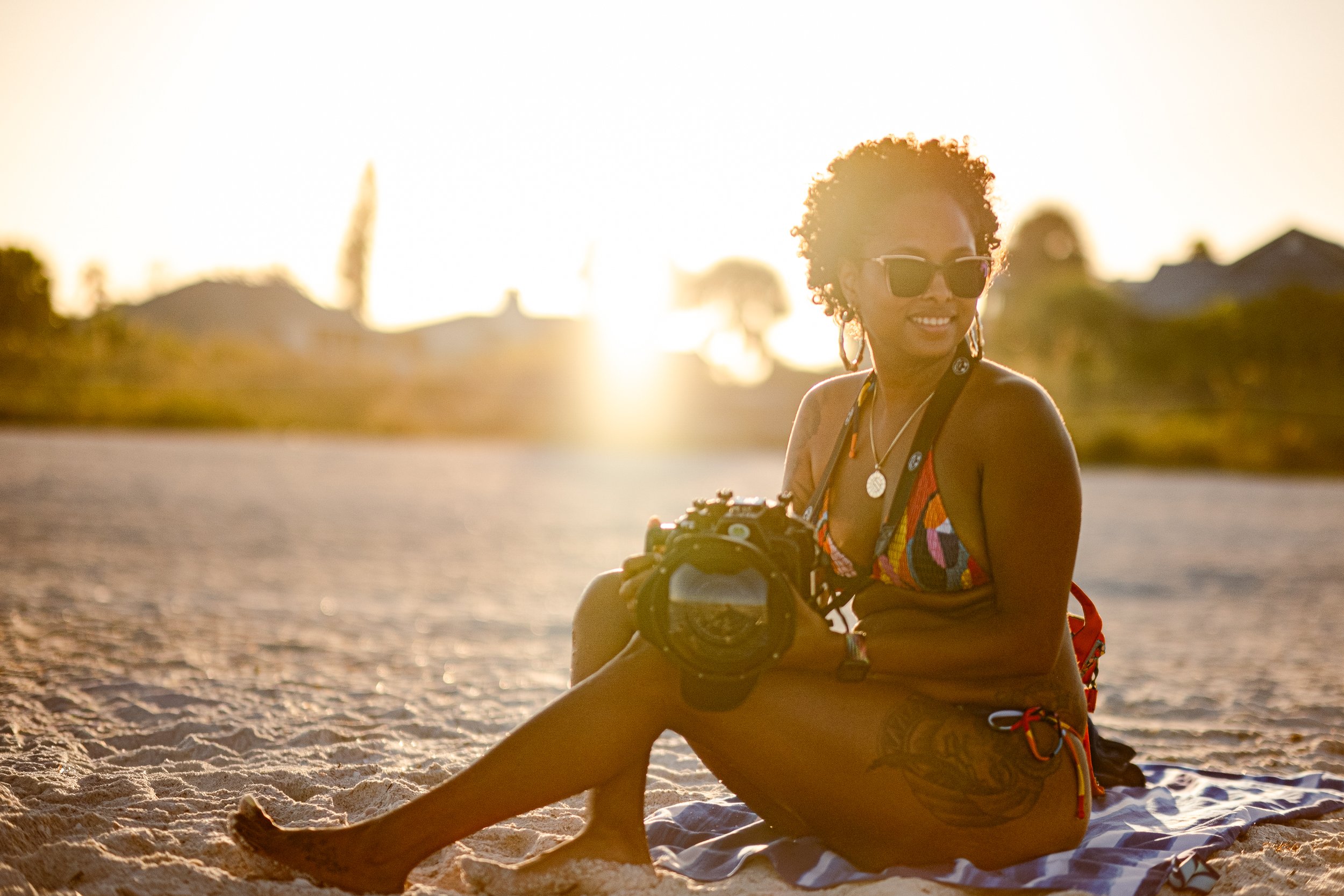 Woman sitting on sandy beach at sunset, wearing sunglasses, colorful tank top, holding camera, smiling.