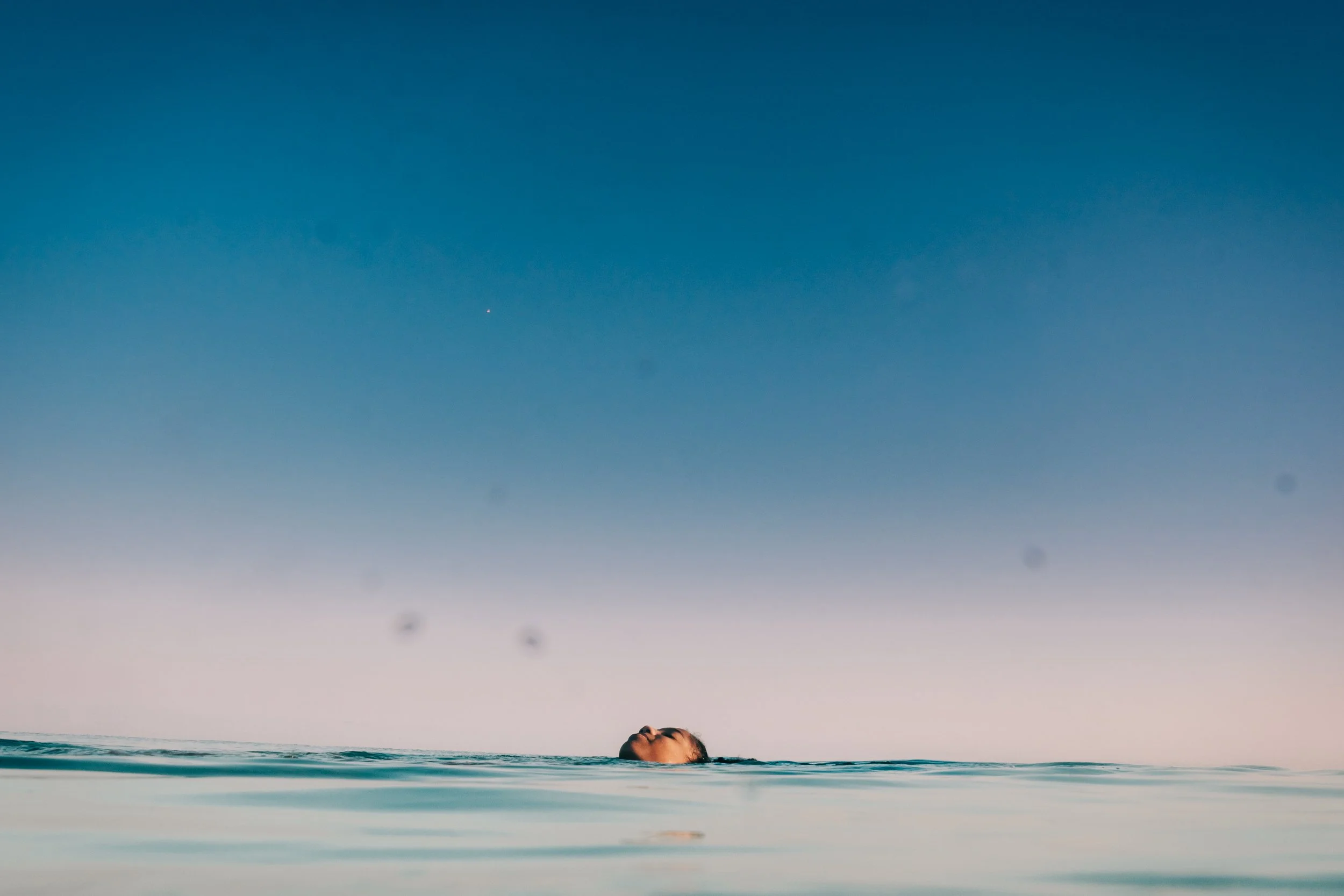 A person swimming in the ocean with only their head visible above the water, under a clear blue sky.