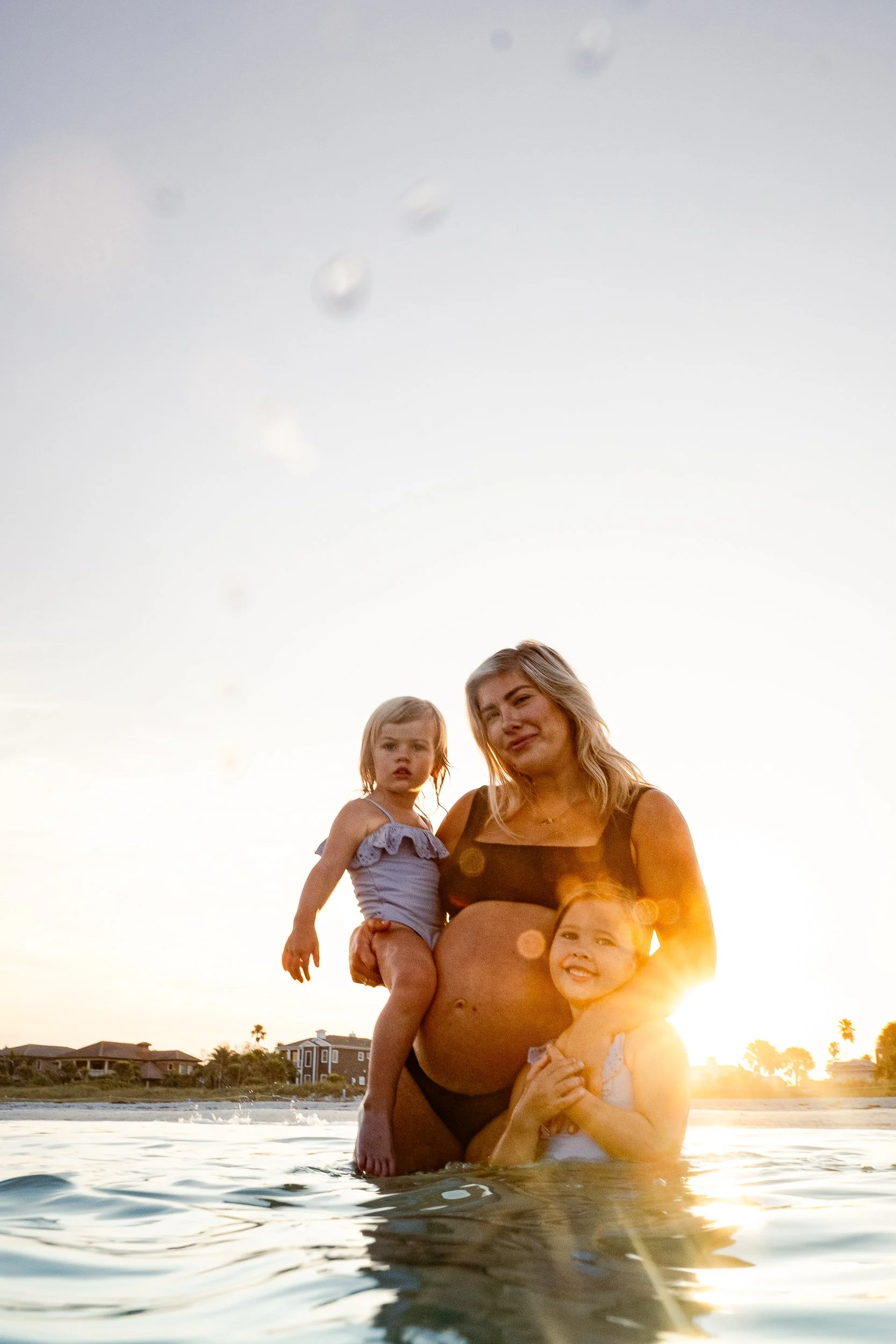 A woman carrying three young children in the water at sunset, with houses and trees in the background.