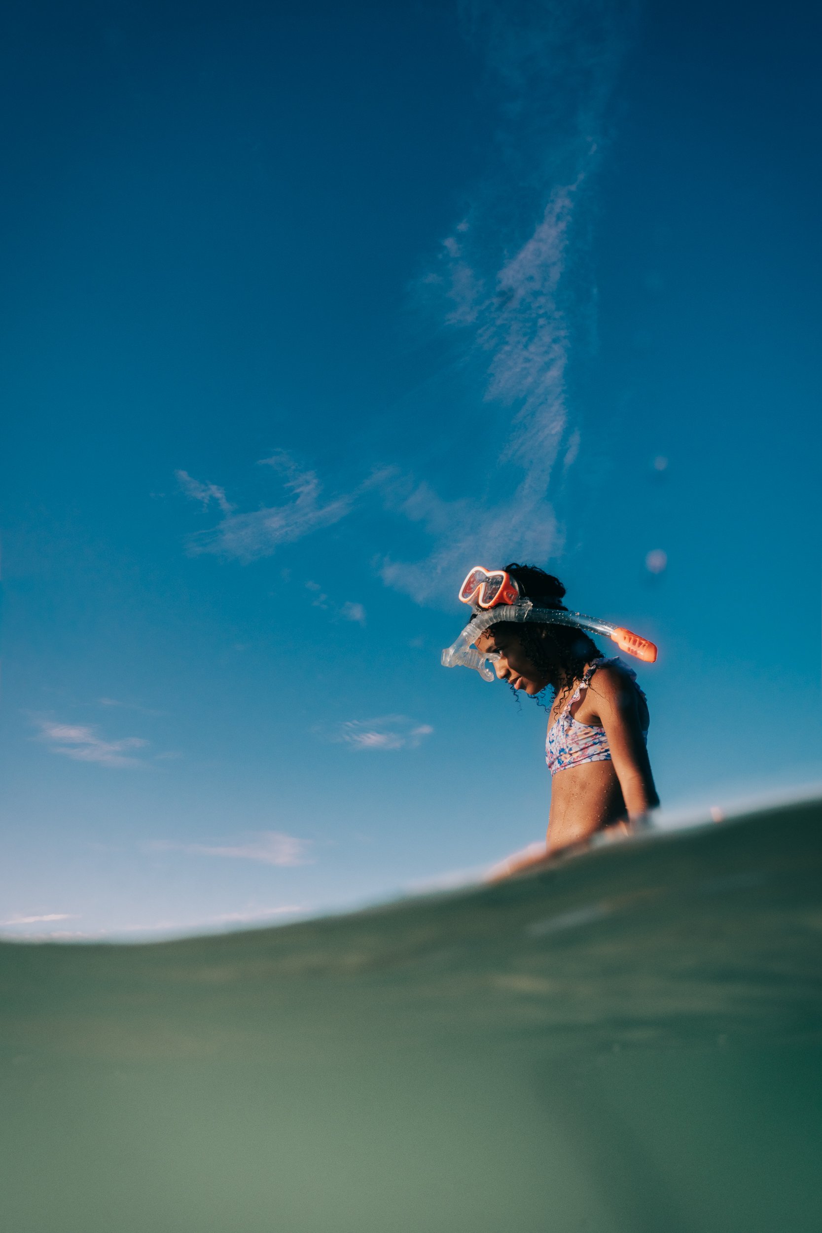 A young woman wearing a bikini and snorkeling gear stands in the ocean, smiling, with a clear blue sky and some clouds overhead, and the water surface visible in the foreground.