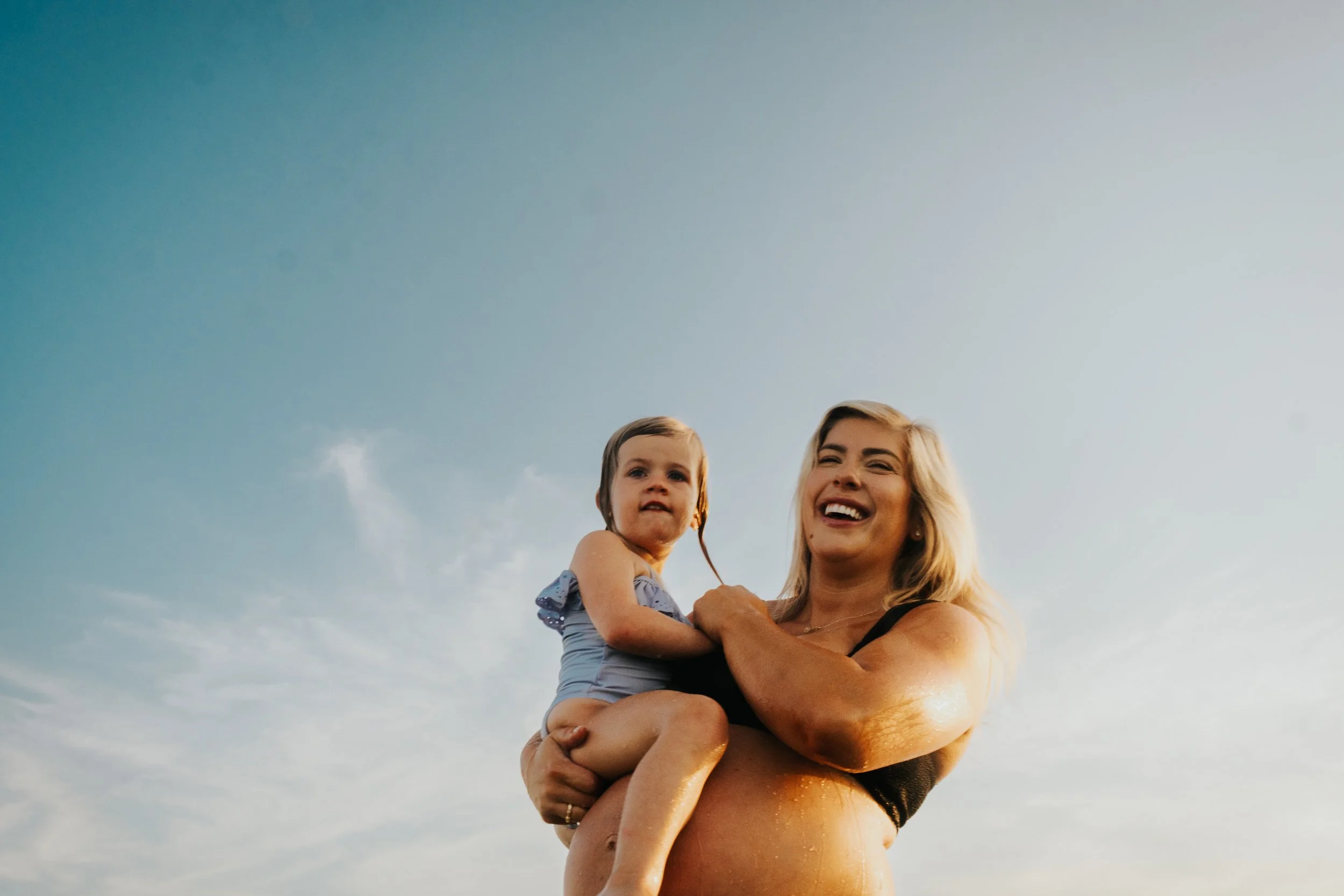 A woman with blonde hair smiling and holding a young girl with brown hair, wearing a blue dress, against a clear blue sky at sunset.