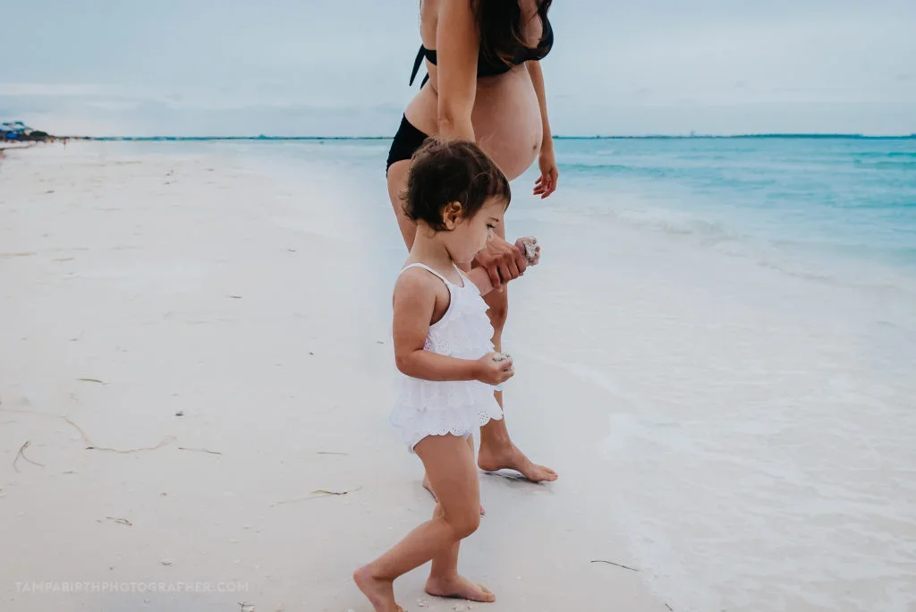 A young girl and pregnant woman walk along the beach, with their feet in the shallow water.