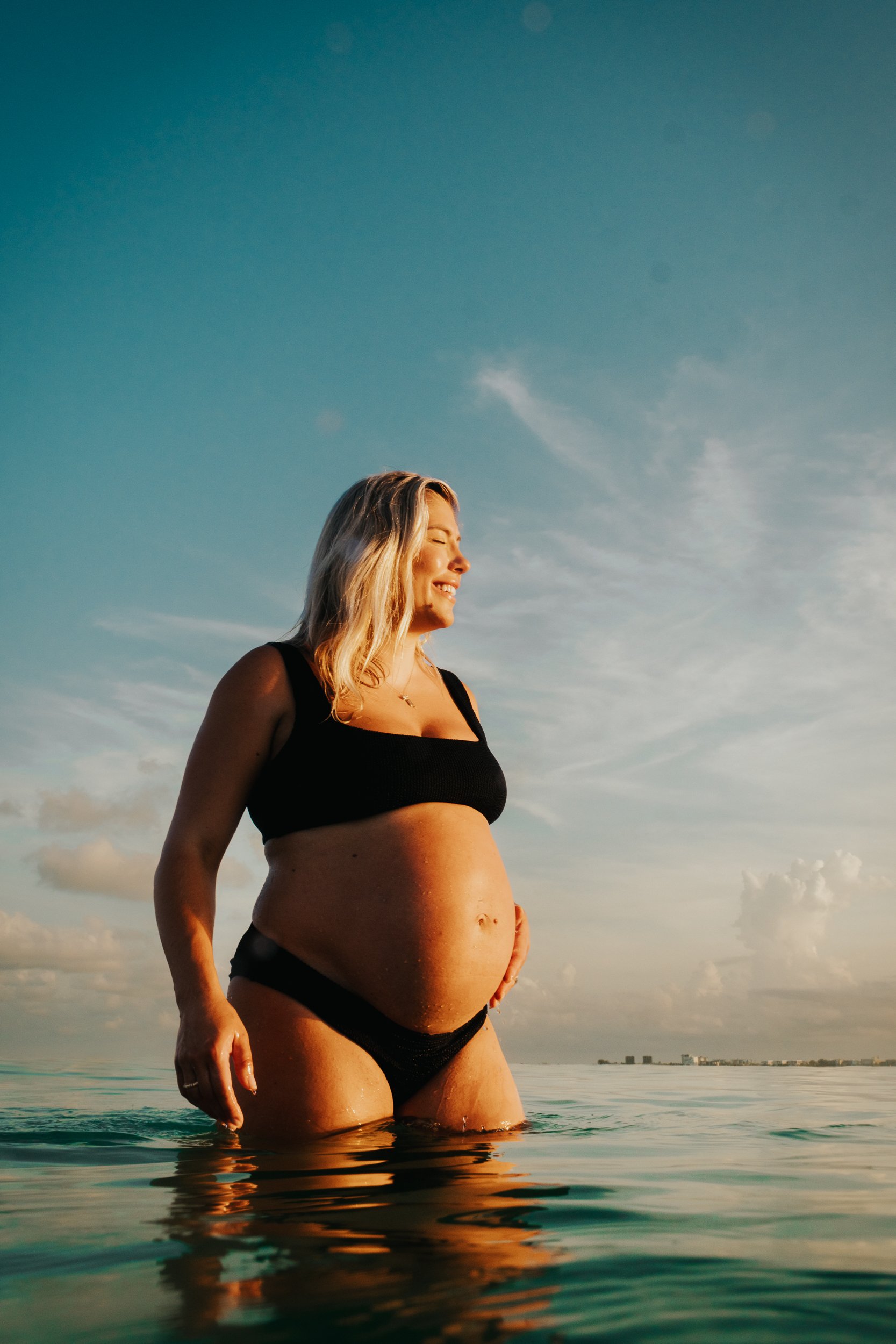 Pregnant woman standing partially submerged in water at the beach, smiling with eyes closed under a sky with some clouds.