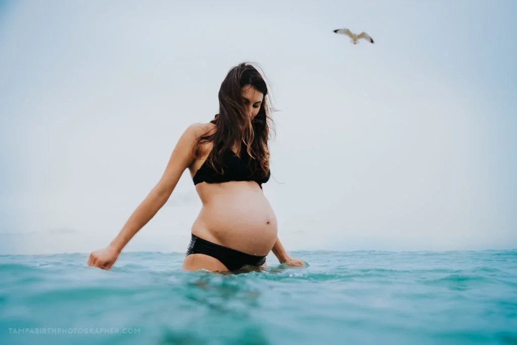 Pregnant woman standing in the water at the beach with a seagull flying overhead.