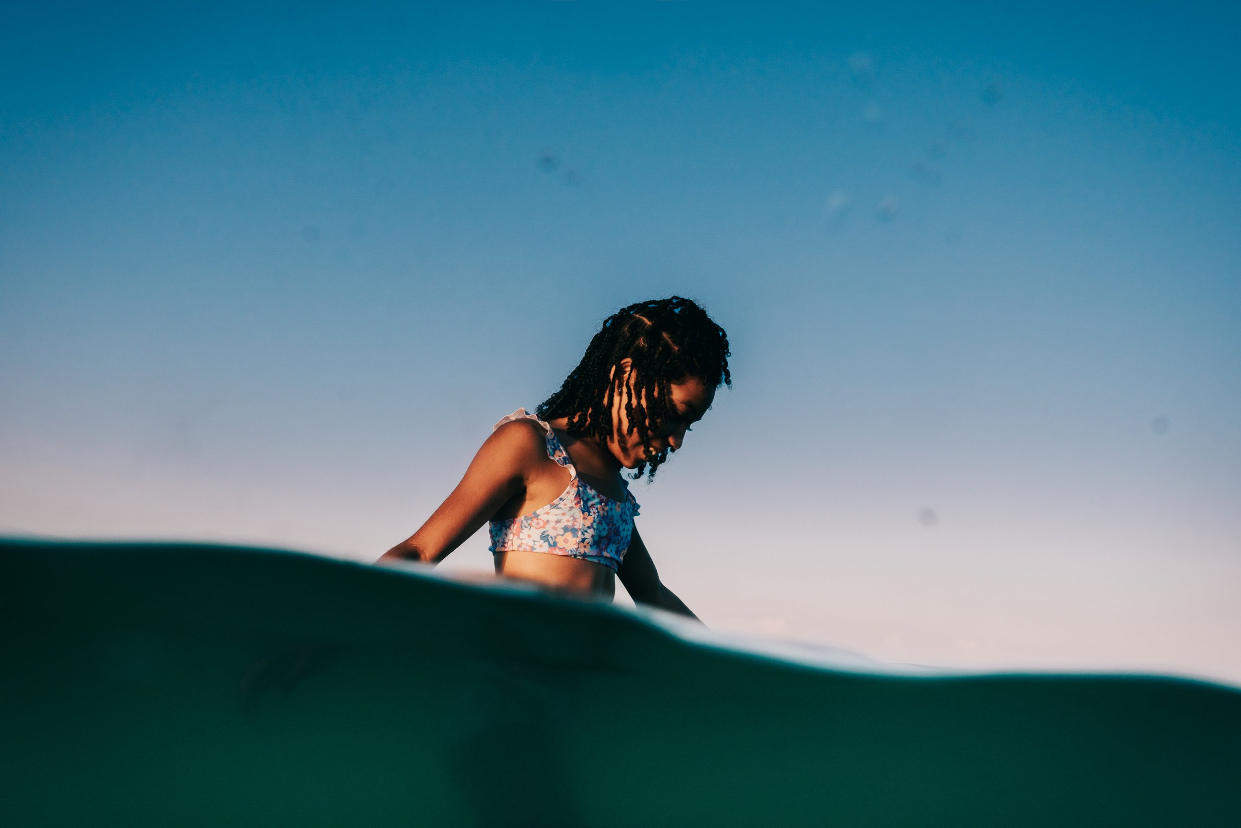A young girl in a floral top looking downward, against a clear blue sky.