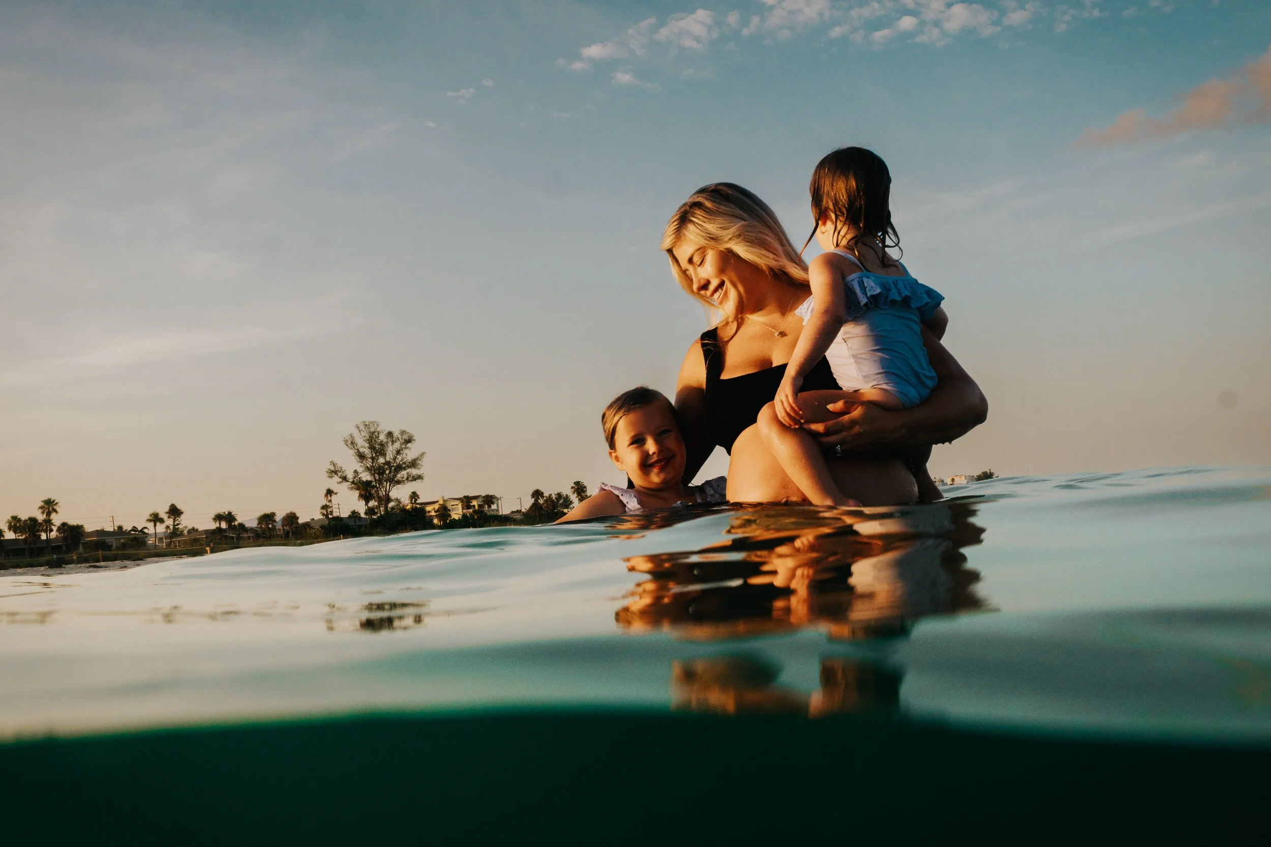 A woman and two young girls enjoying a moment in the water at sunset, with the sky and distant shoreline visible in the background.