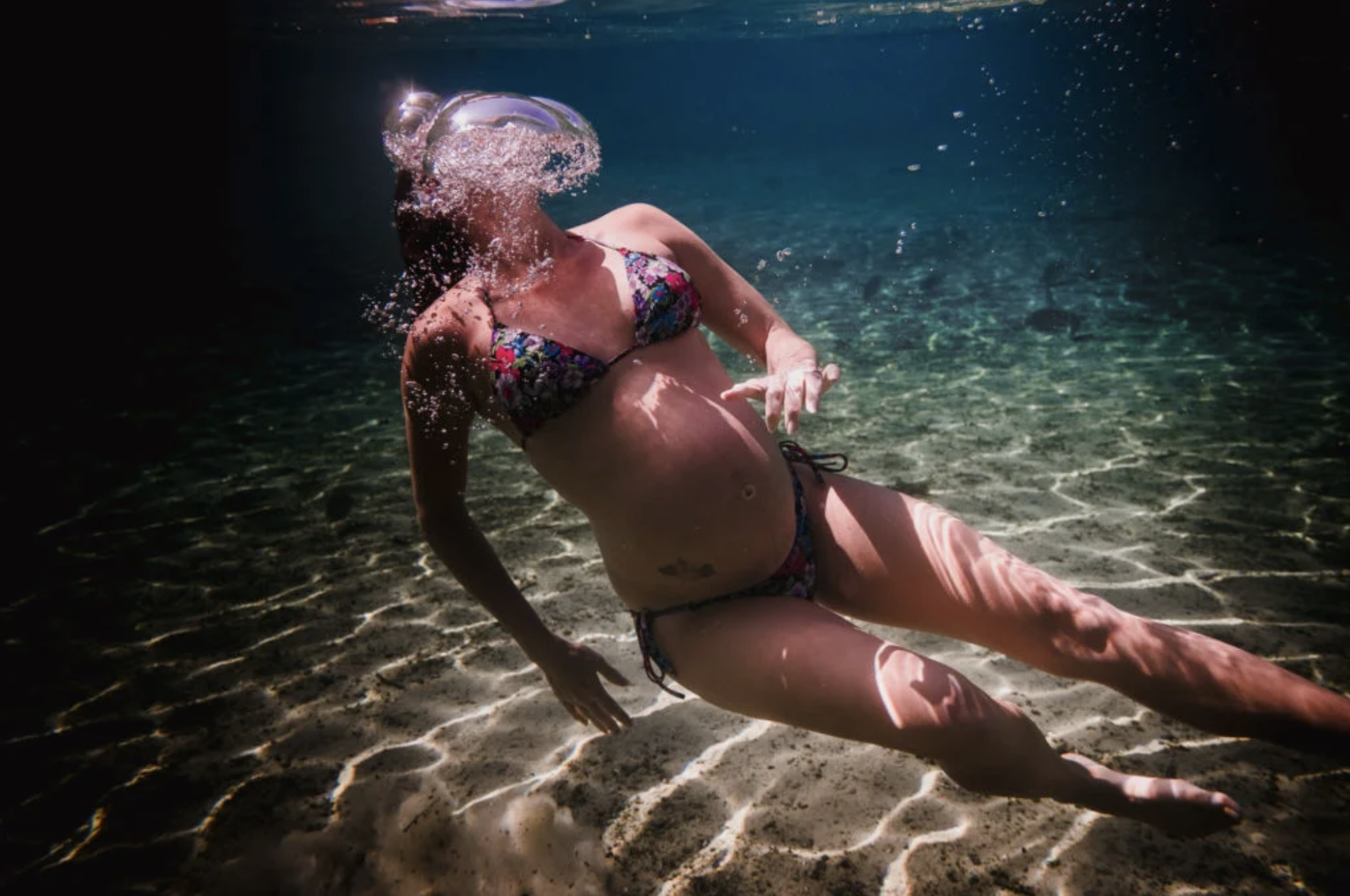A woman in floral bikini underwater, with hair and bubbles near her head, sitting on the sandy ocean floor.