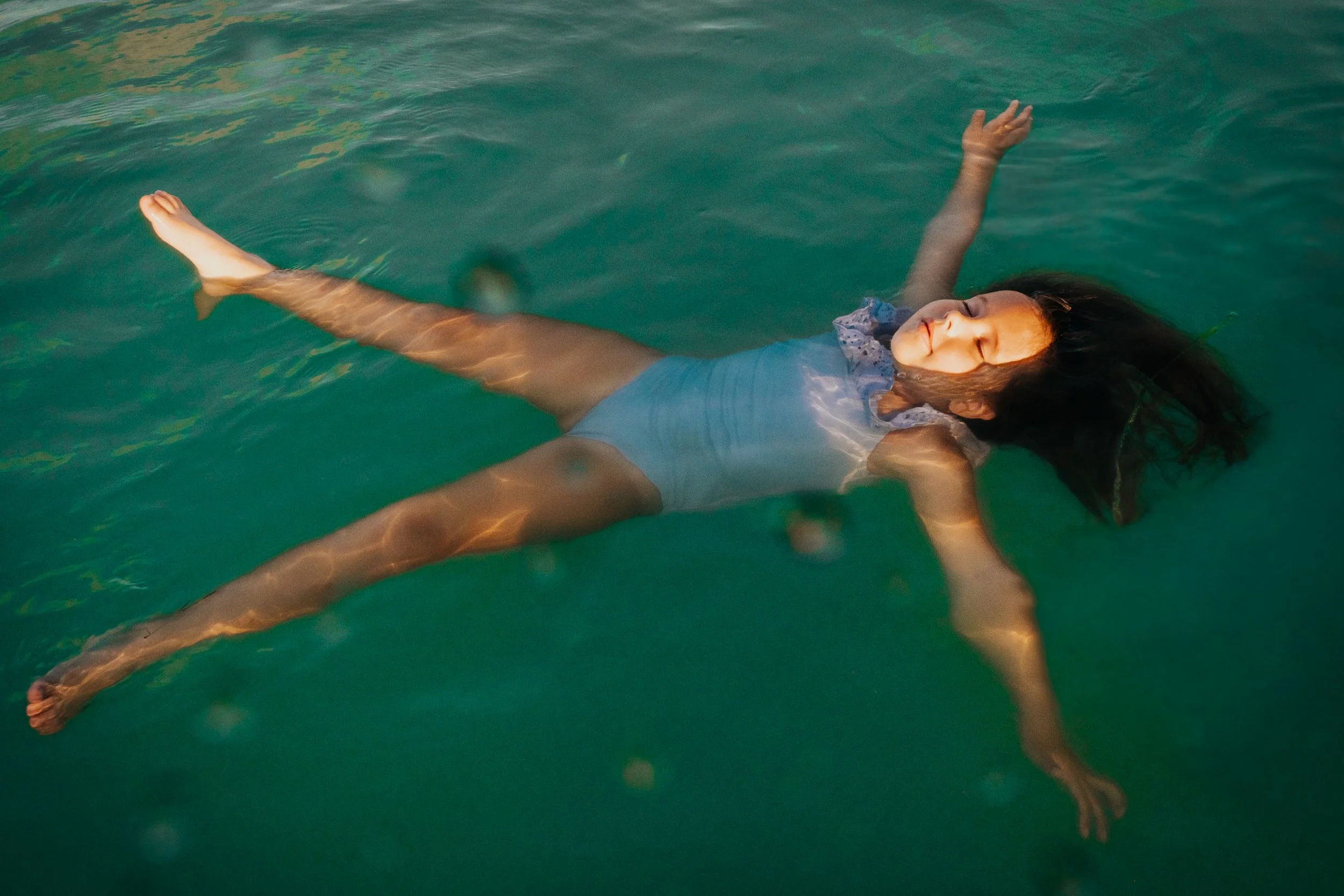 A young girl with dark hair floating on her back in a swimming pool, smiling with eyes closed and arms outstretched.