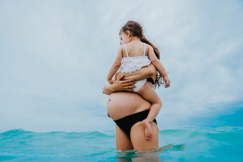 Person holding a young girl in the ocean with the sky and water in the background.
