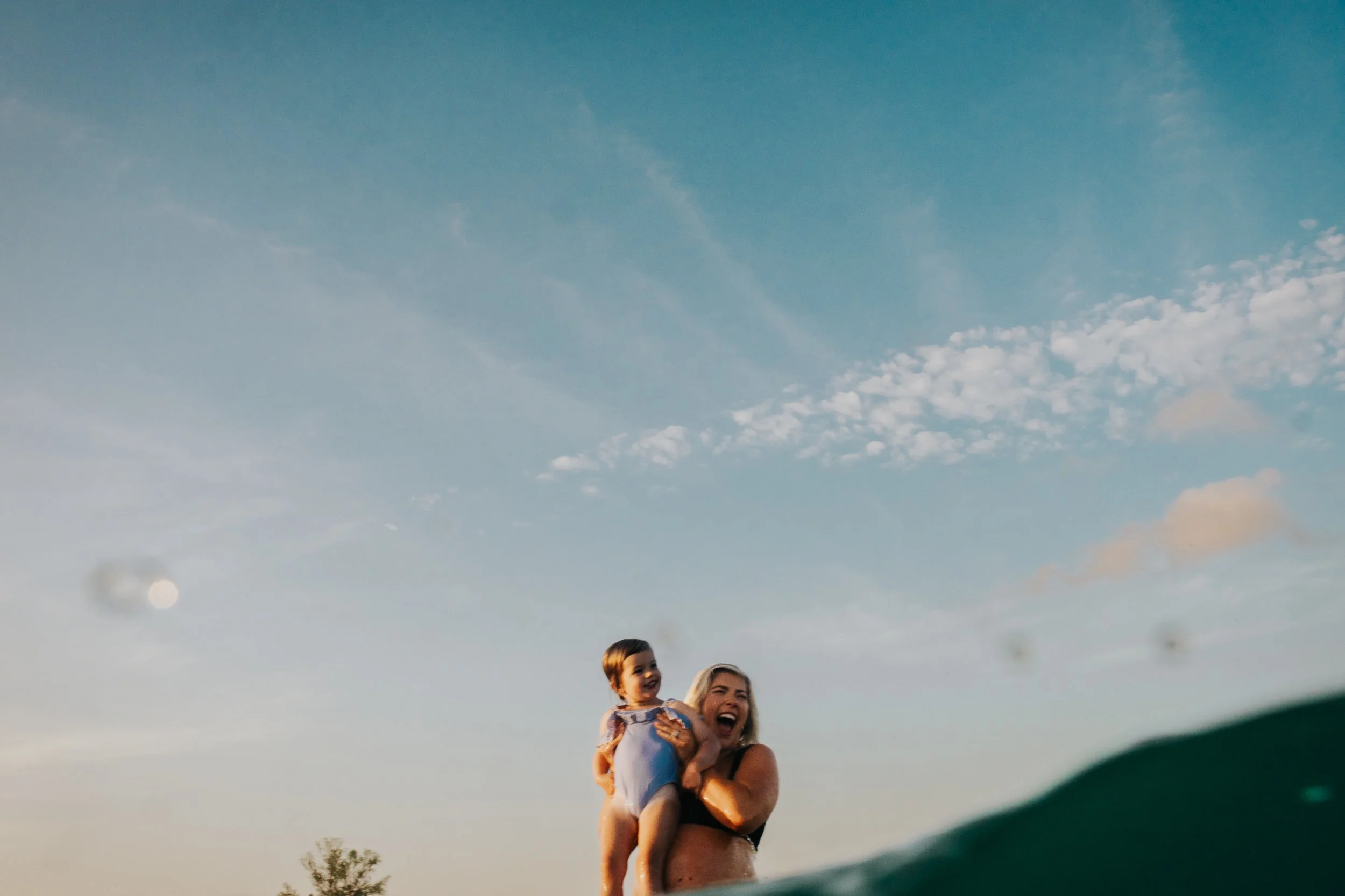 A woman is holding a young girl on her shoulders outdoors during daylight, with a bright sky and some clouds in the background. Both are smiling and appear to be enjoying themselves.