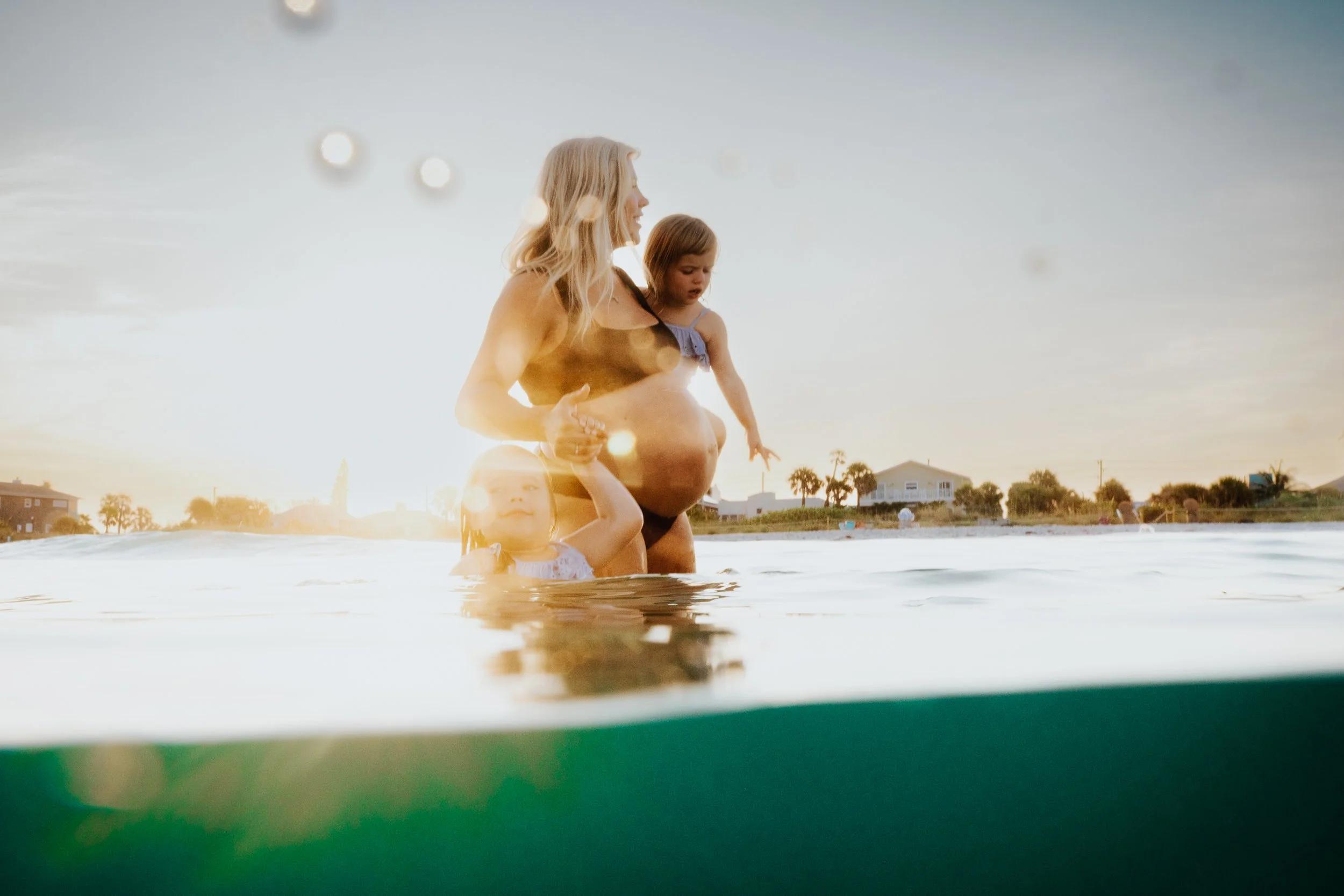 A woman and two young girls in a body of water at sunset, with houses and palm trees in the background.