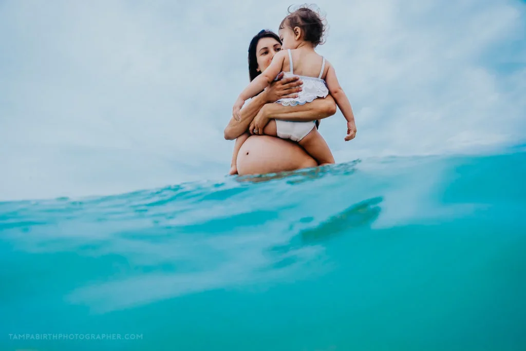 A woman is holding a young girl in her arms while standing in the ocean, with the water reaching up to her waist. The woman appears to be pregnant, and both are looking at each other.