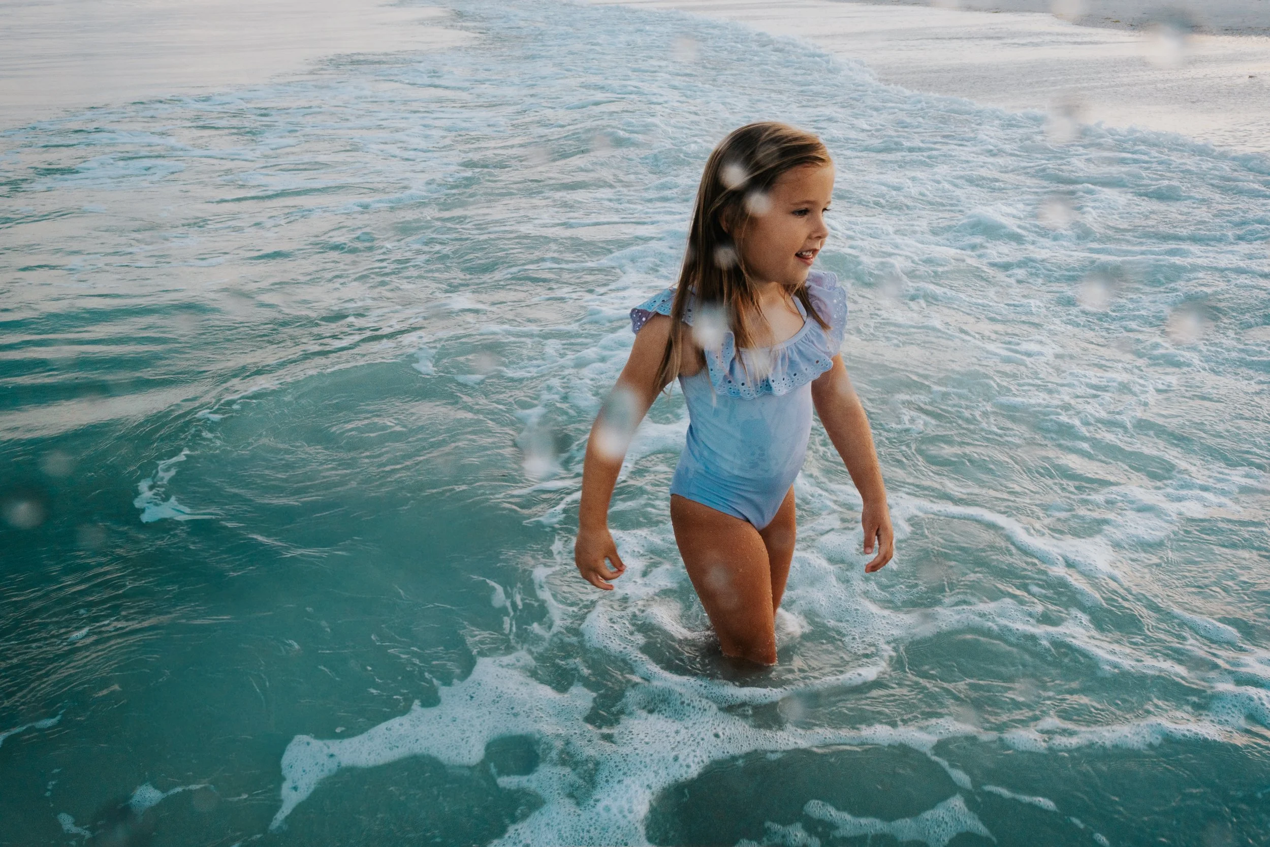 A young girl in a light blue swimsuit standing in the ocean, with water splashing around her, and a trail of white foam behind her as she moves forward in the sea.