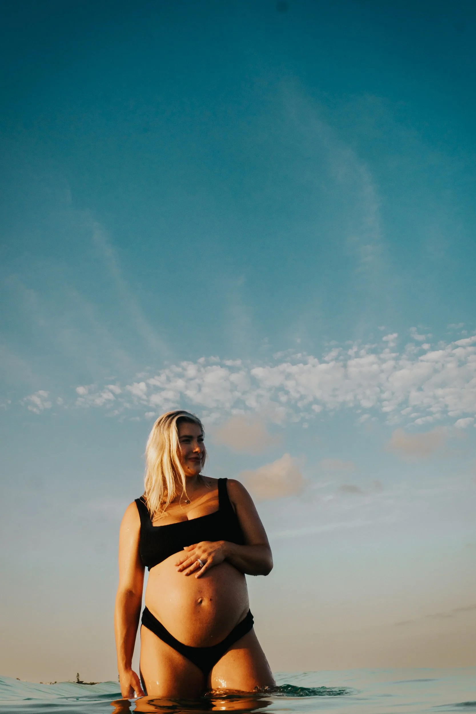 A woman with blonde hair in a black bikini stands waist-deep in water, with a sky filled with scattered clouds overhead during sunset.