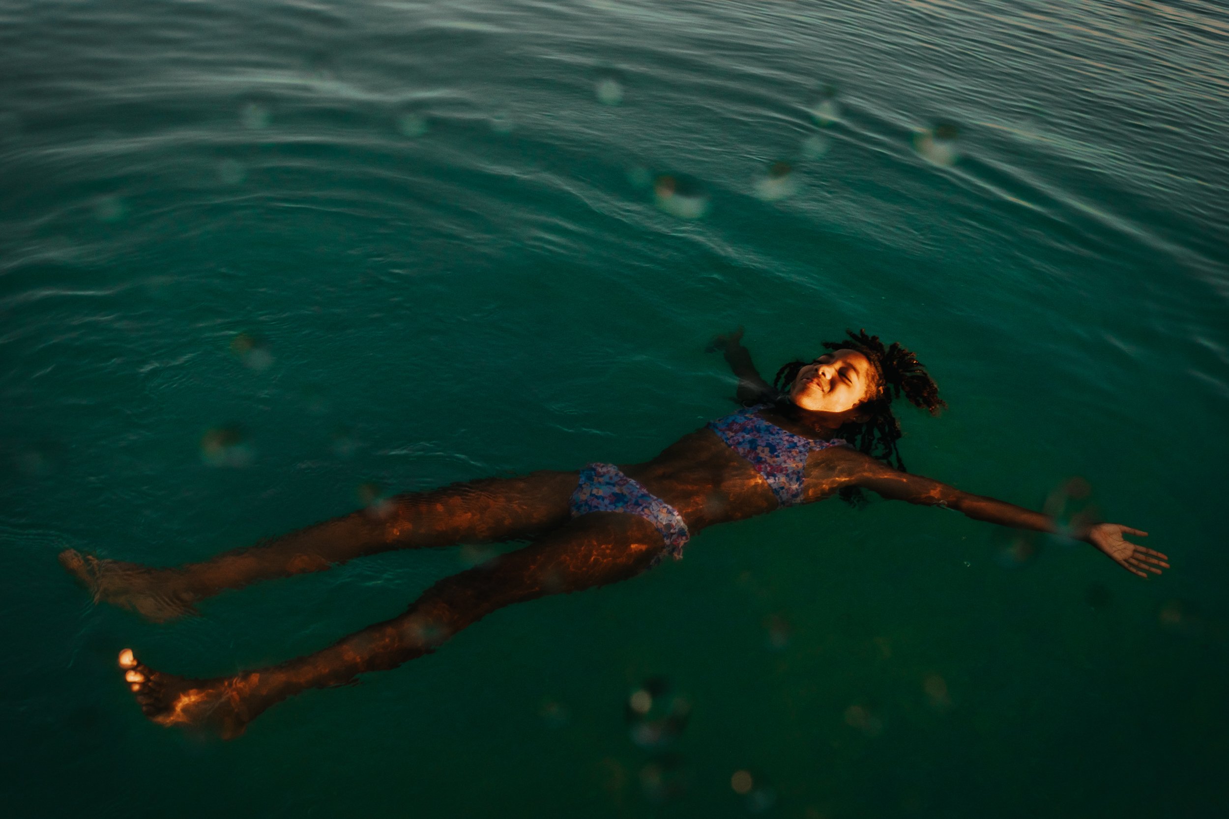 A woman with dreadlocks is floating on her back in a body of water, smiling with her eyes closed, wearing a colorful swimsuit.
