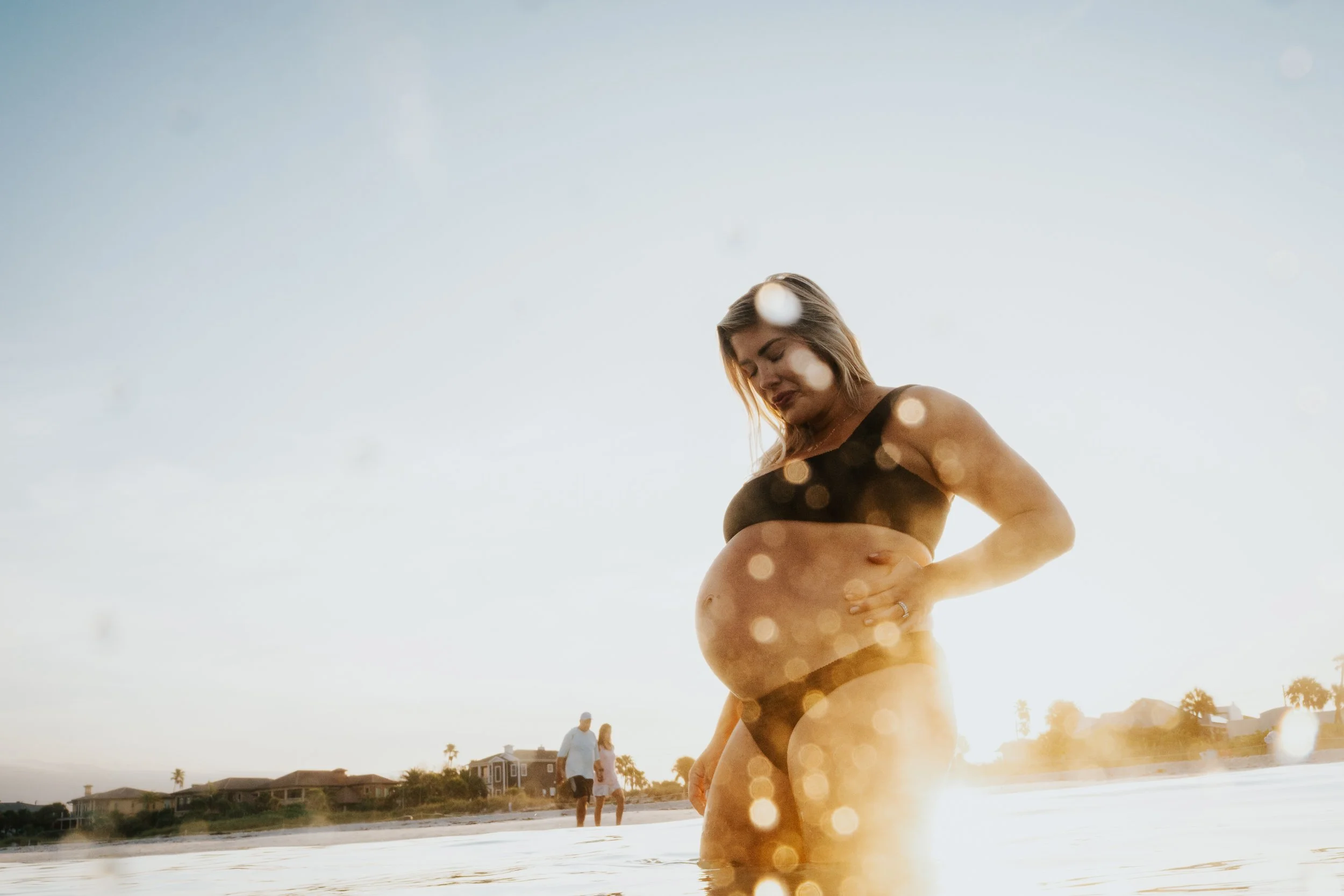 A pregnant woman standing on the beach during sunset, with people walking in the background and palm trees visible.