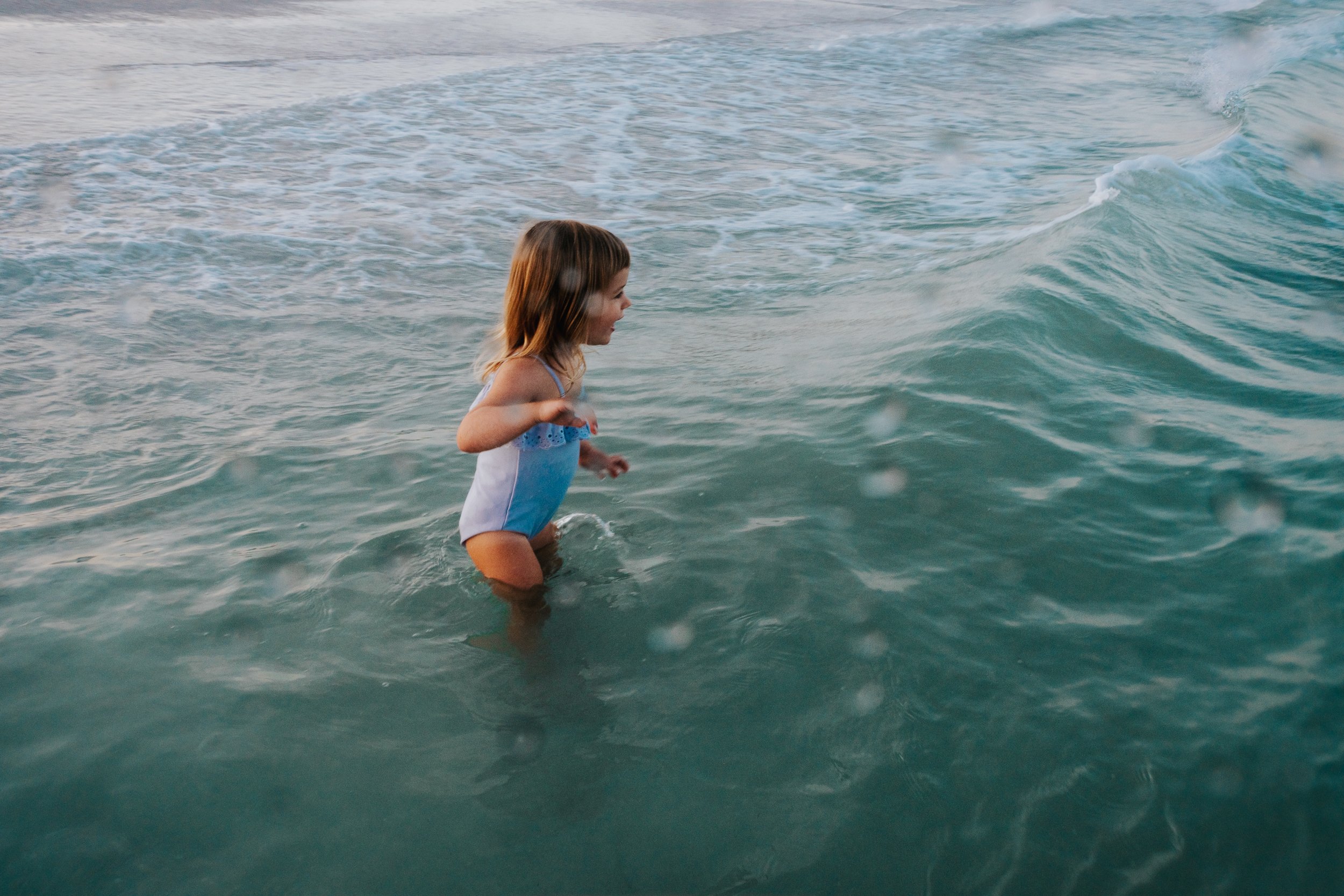 A young girl in a blue swimsuit standing in the water at the beach, smiling as she faces to the right with small waves around her.