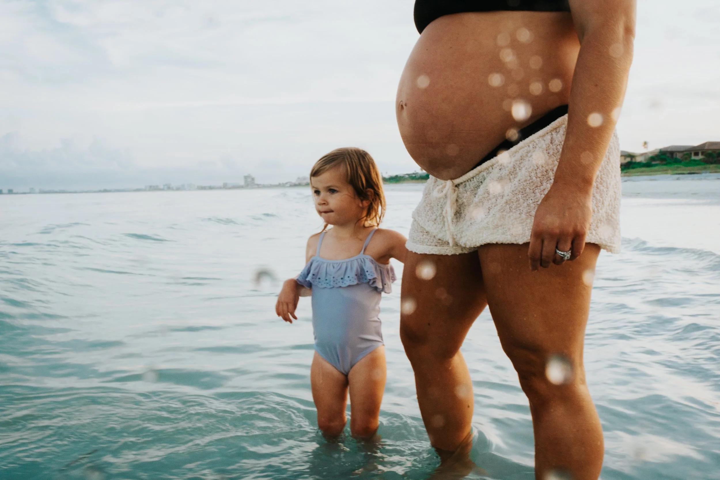 A woman and a young girl are standing in the ocean at the beach, with the woman holding the girl’s hand. The woman is wearing cozy, beige shorts, and the girl is wearing a light blue swimsuit with ruffles. The background shows an overcast sky and bui