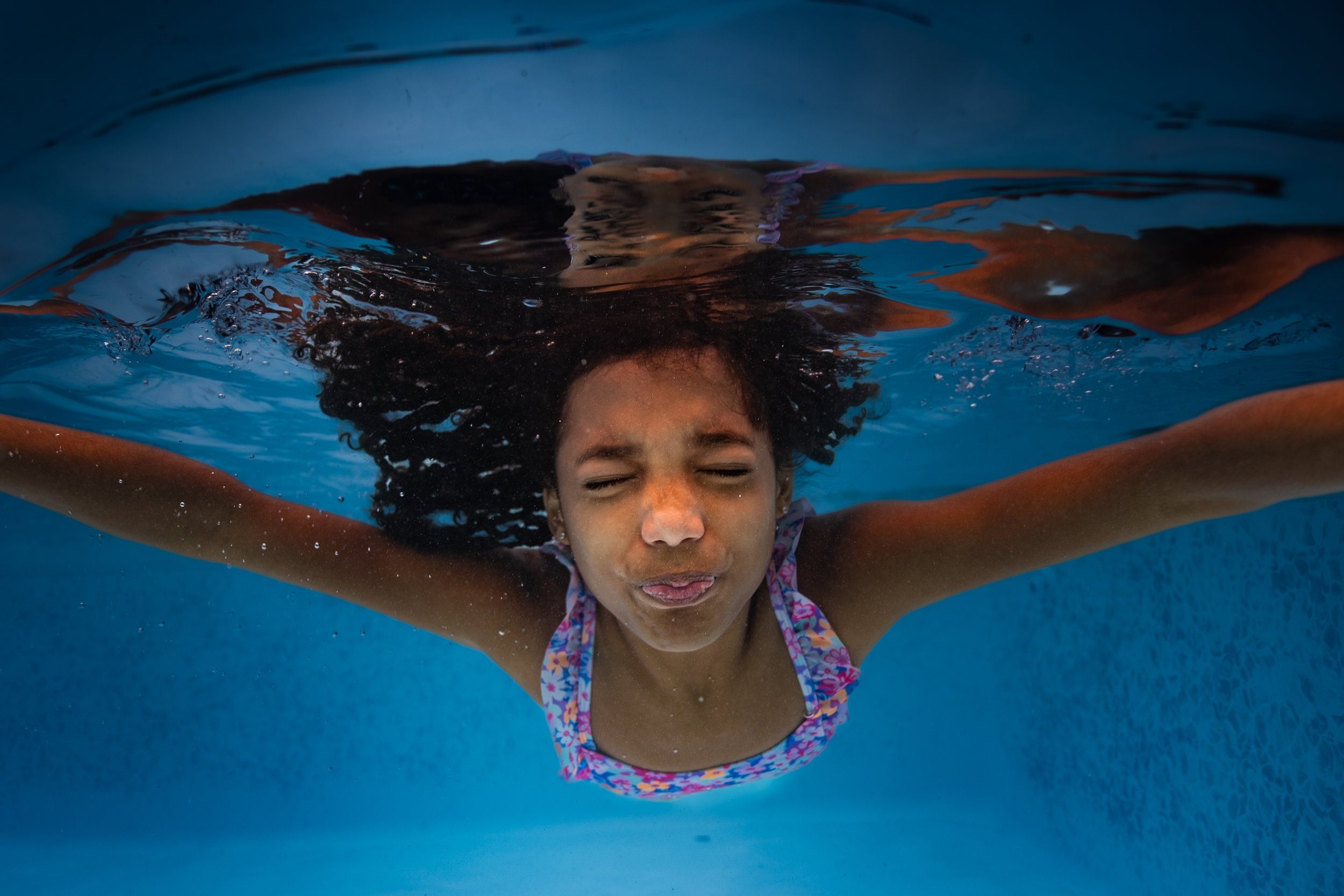 Young girl swimming underwater with her eyes closed, arms outstretched, and wearing a colorful swimsuit.