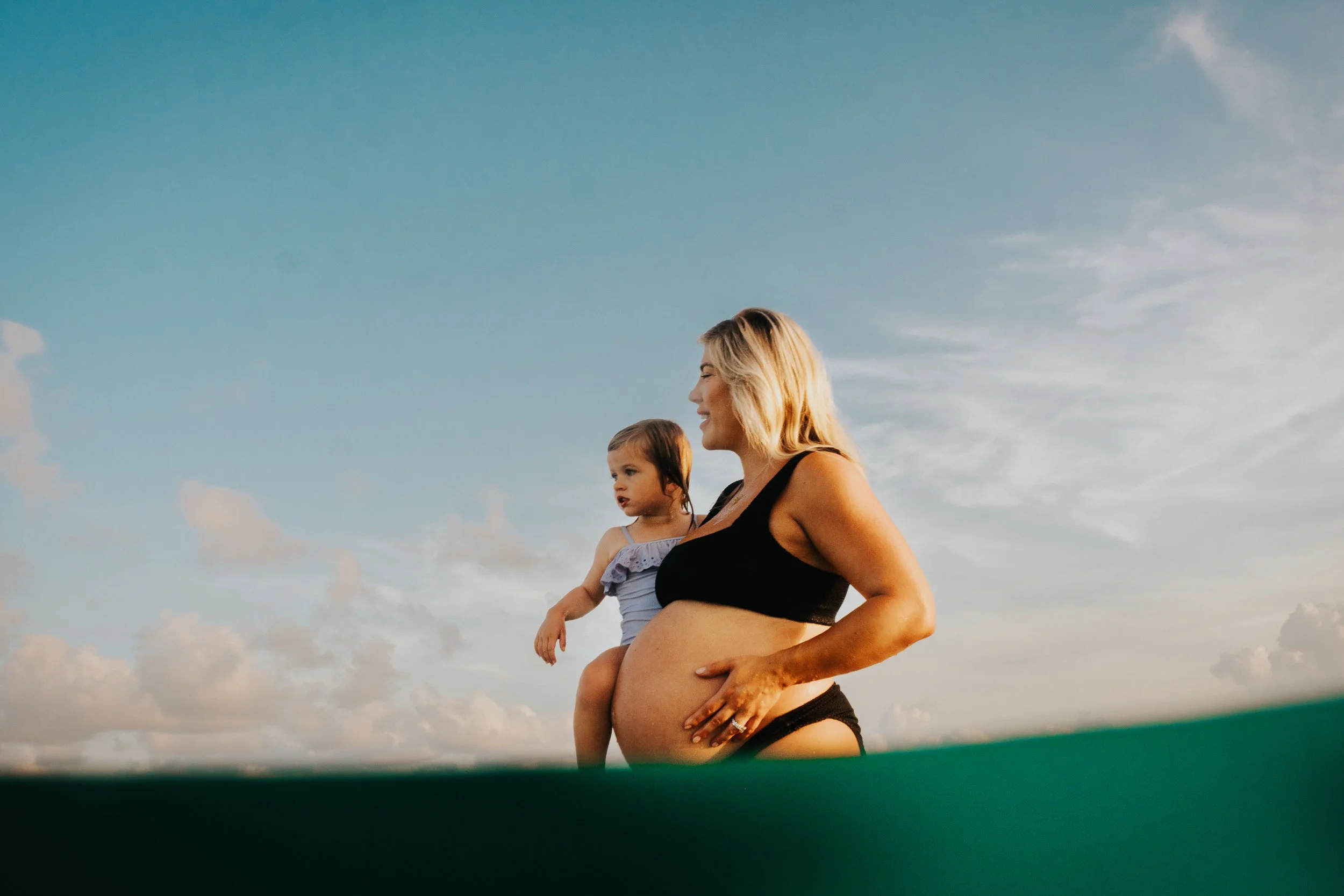 A pregnant woman holding a young girl in her arms outdoors under a blue sky with some clouds.